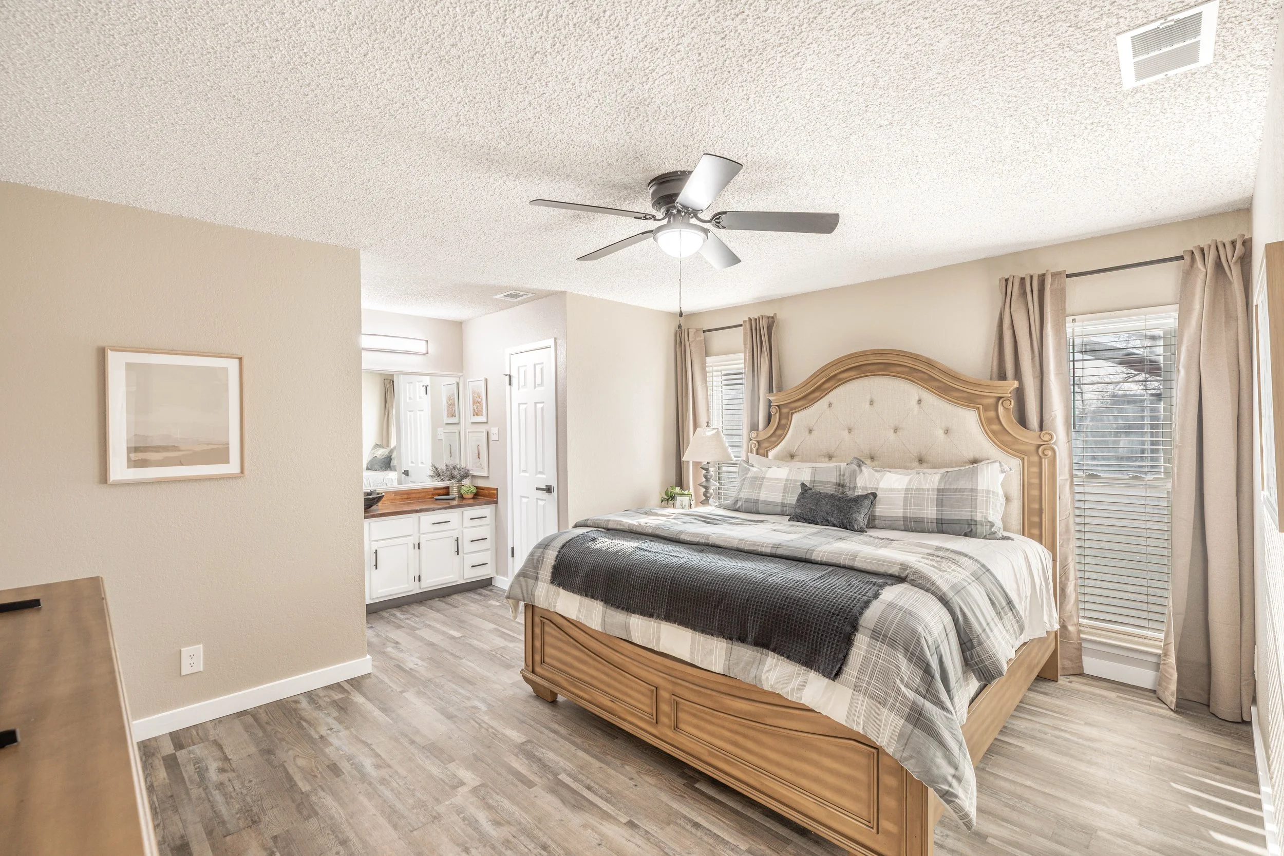 A bedroom with a wooden bed frame, beige headboard, plaid bedding, and windows with curtains. There are two white doors, a ceiling fan, and framed artwork on the walls.