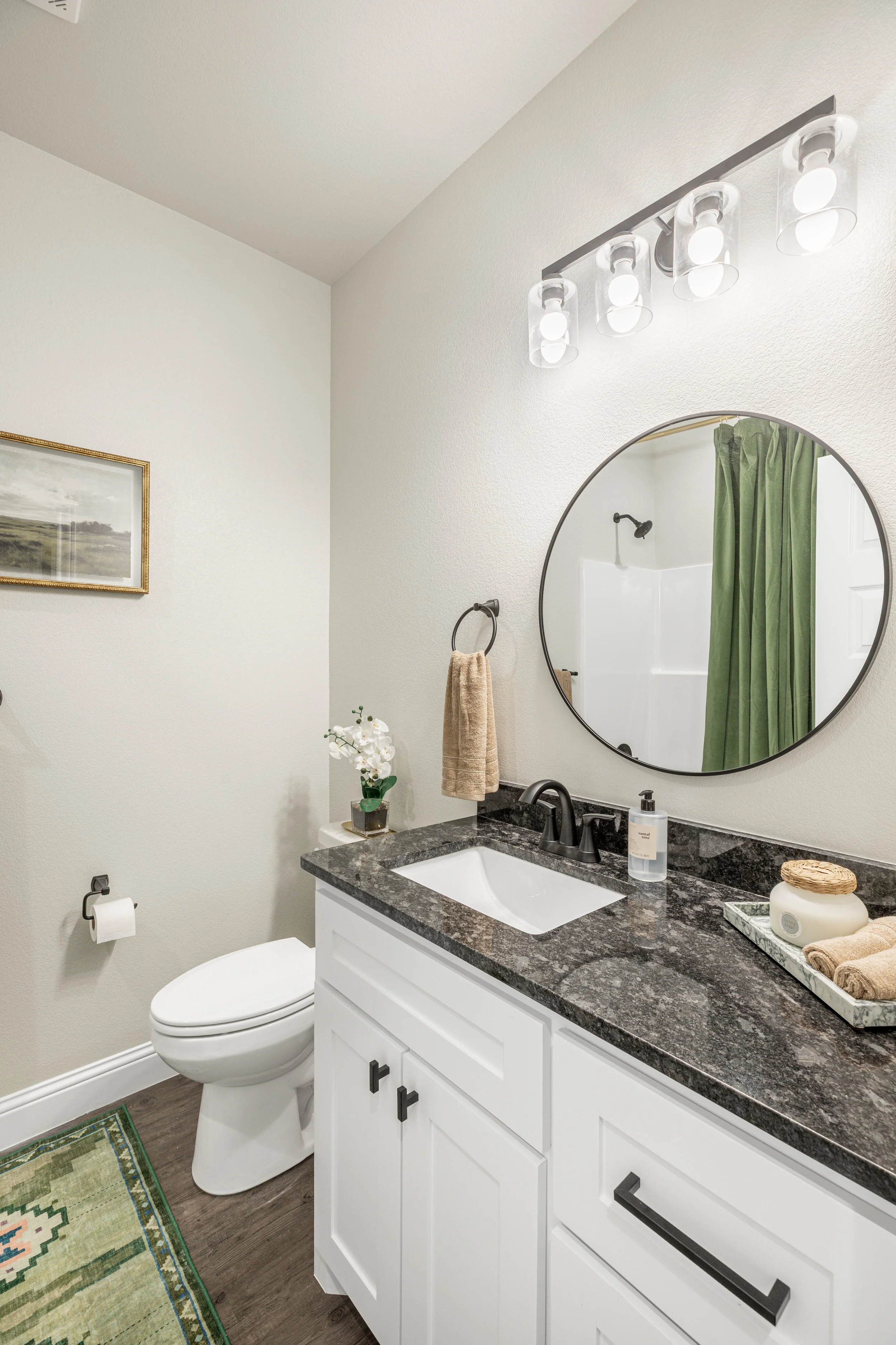 A bathroom with a white vanity, black granite countertop, round mirror, towels, a plant, and a toilet, with wall art and a shower with green curtain.