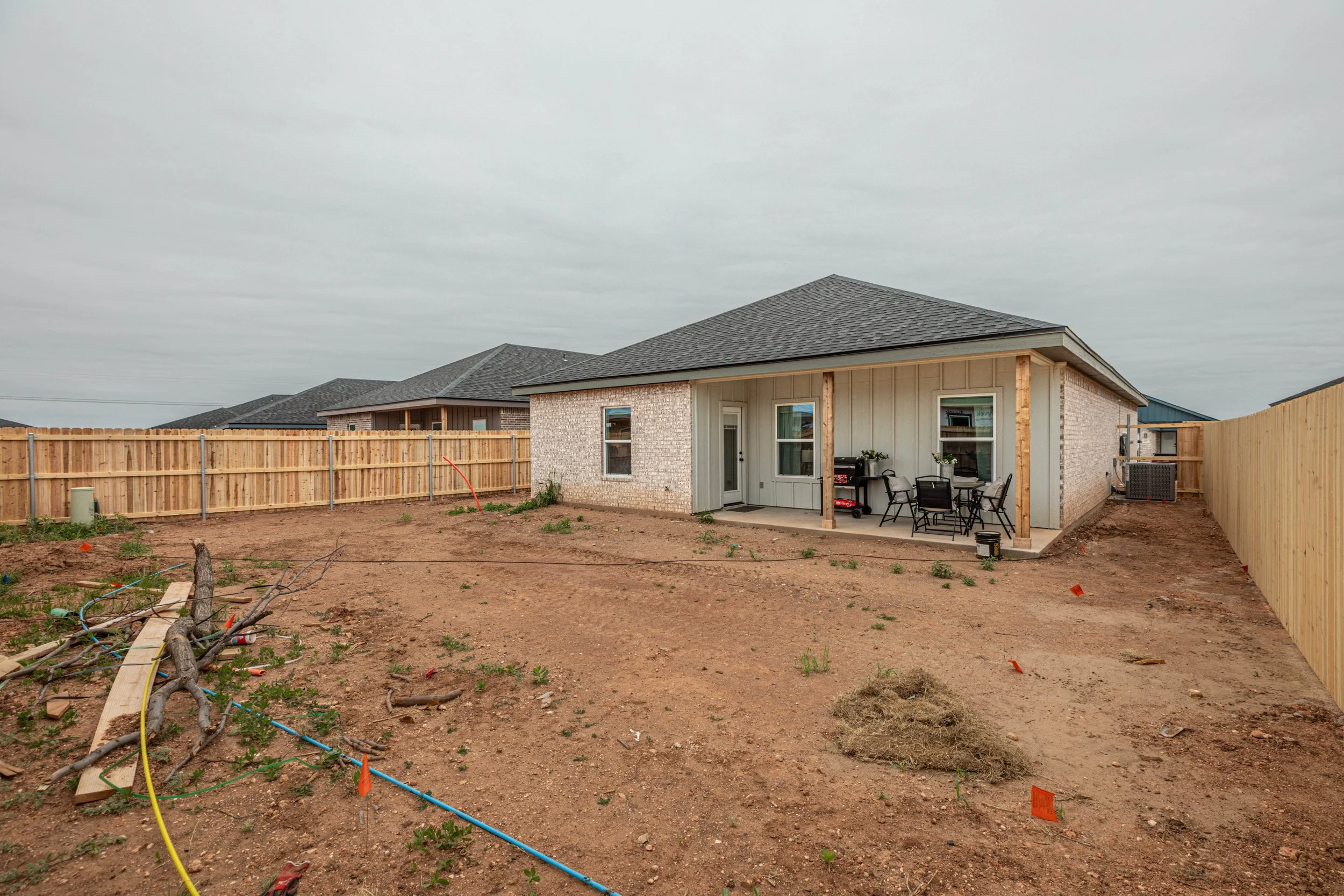 Backyard of a house under construction with fenced yard, patio furniture, and an unfinished yard with soil and construction materials.