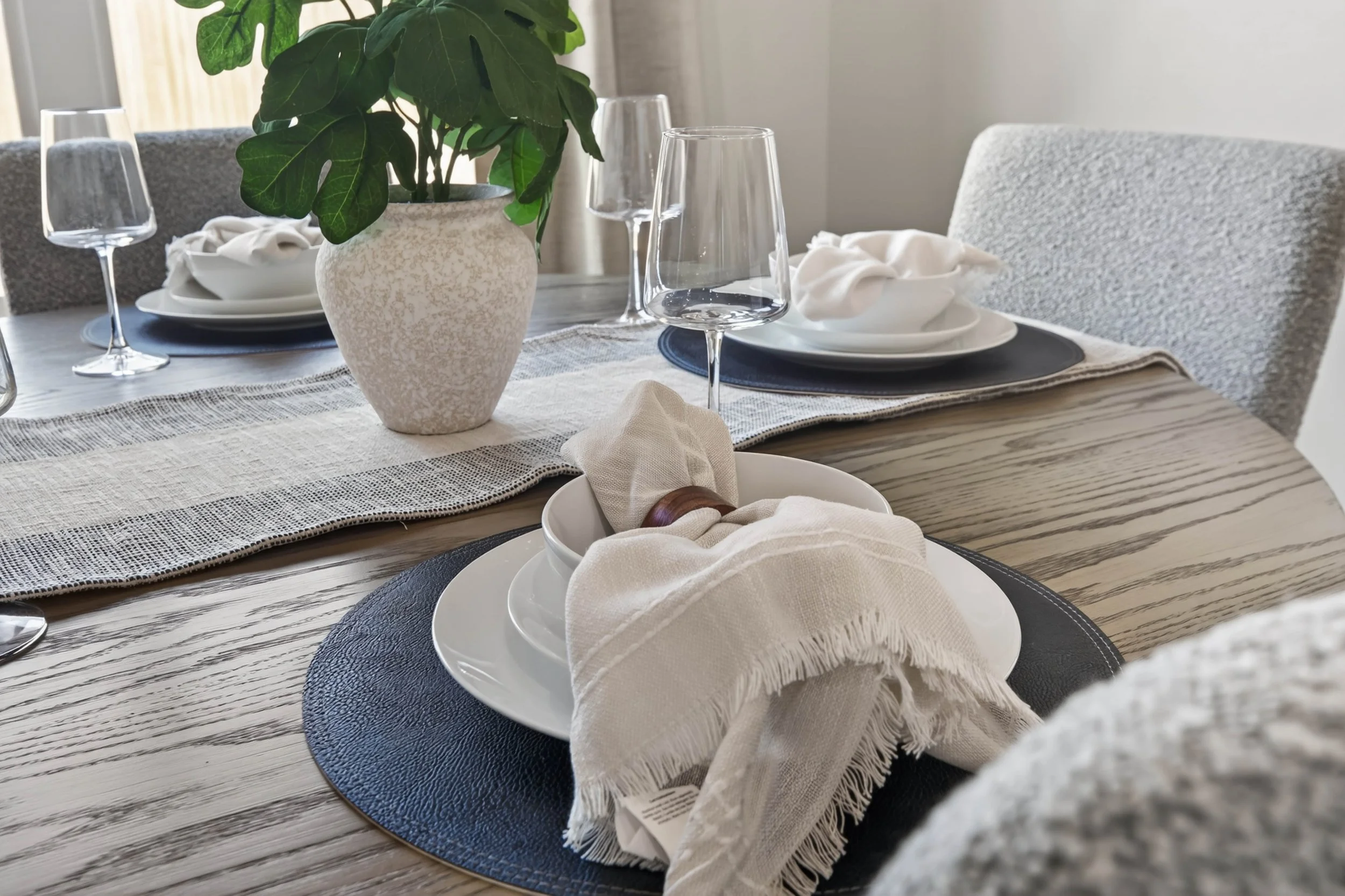 A dining table decorated with a potted plant, wine glasses, a stack of plates with napkins, and a textured table runner. The chairs are upholstered in a light-colored fabric, and the setting appears ready for a meal.