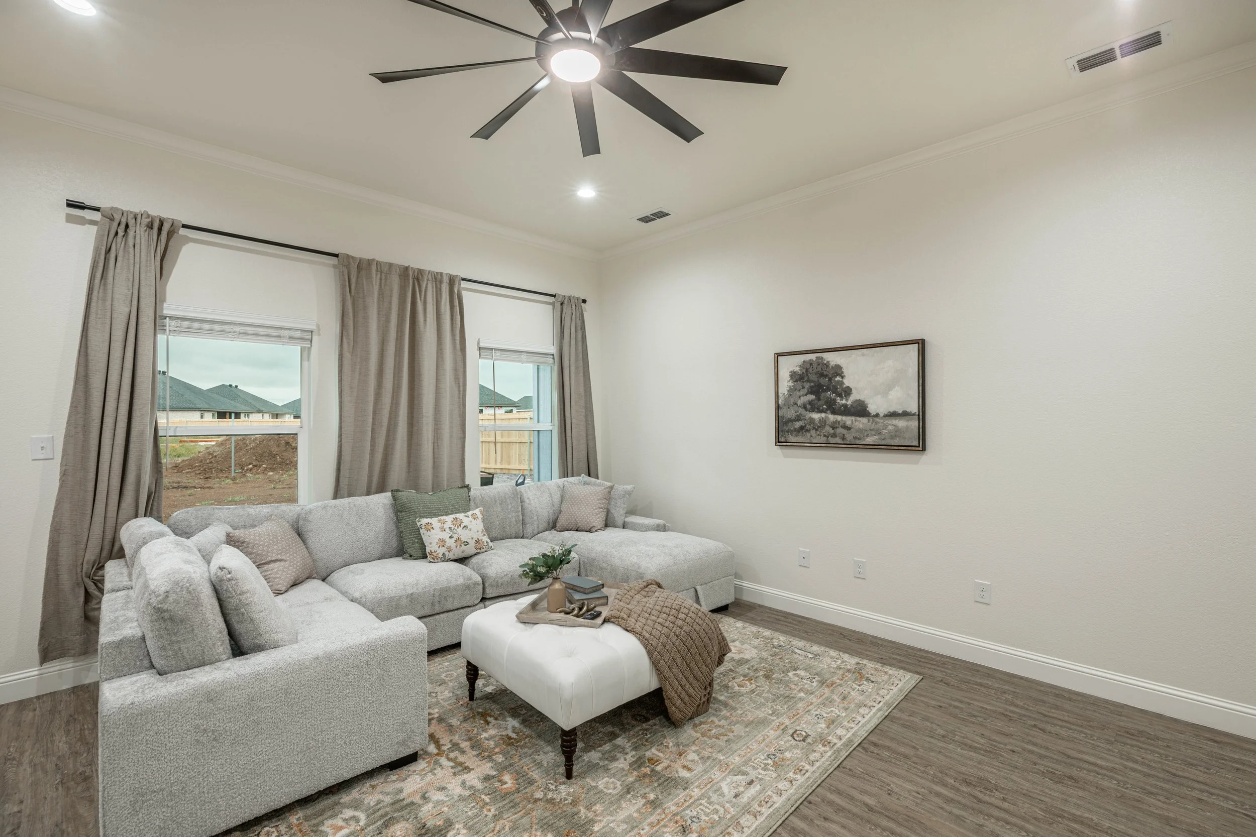 Living room with a gray sectional sofa, decorative pillows, a white ottoman, a patterned area rug, wall art, and large windows with beige curtains. Ceiling fan with dark blades is mounted on the ceiling.