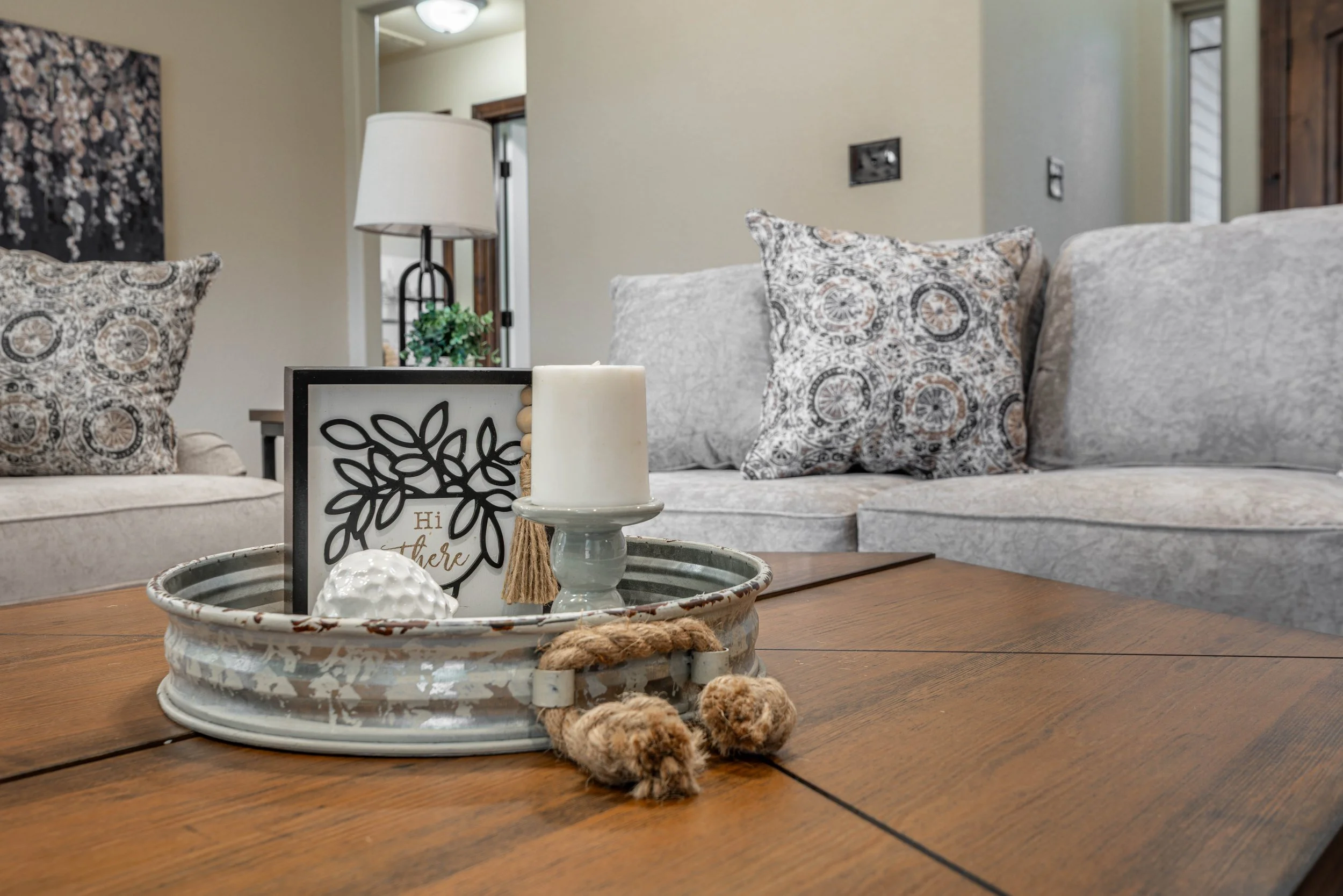 Interior of a living room with a wooden coffee table, decorative tray with candles and a framed quote, a gray sofa with patterned pillows, and a wall with artwork and a window.
