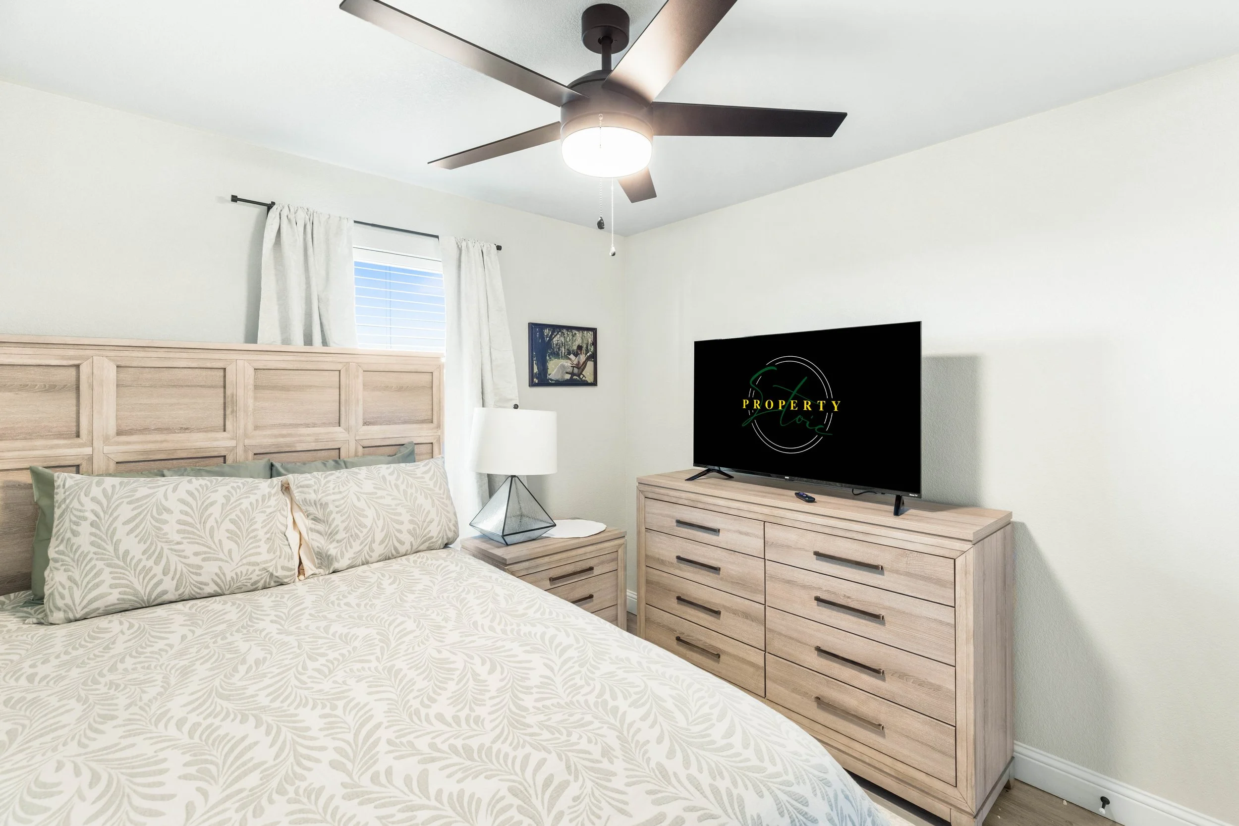 A bedroom with a white bedspread decorated with a leafy pattern, a wooden headboard, a bedside table with a geometric table lamp, a flat-screen TV on a wooden dresser, and a window with white curtains. A ceiling fan with a light is also visible.