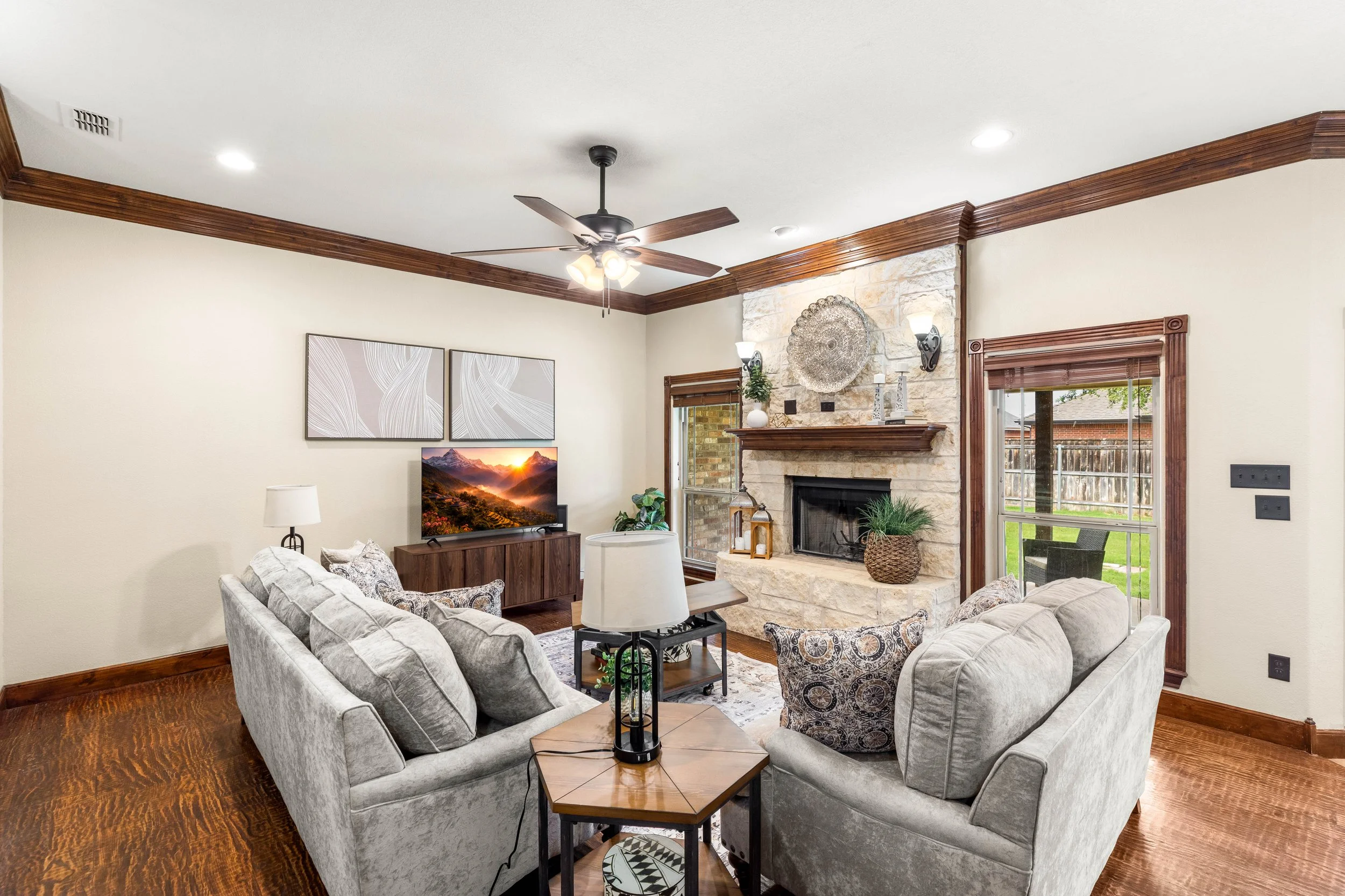 Living room with a beige couch, armchair, and wooden coffee table. Features a stone fireplace, decorative artwork, and a ceiling fan with lights.