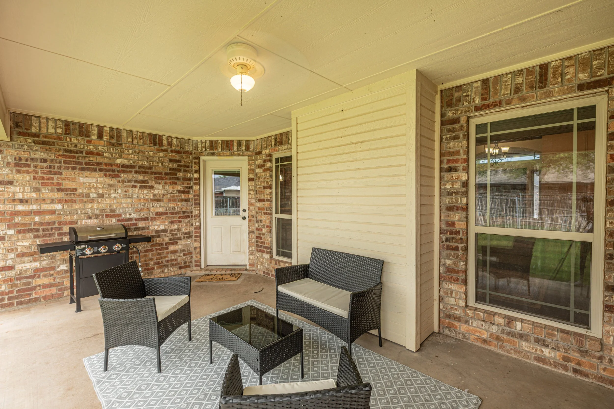 Covered patio area with brick walls, a yellow ceiling, a ceiling fan, large windows, a glass-top table with black wicker chairs, a small rug, and a grill.