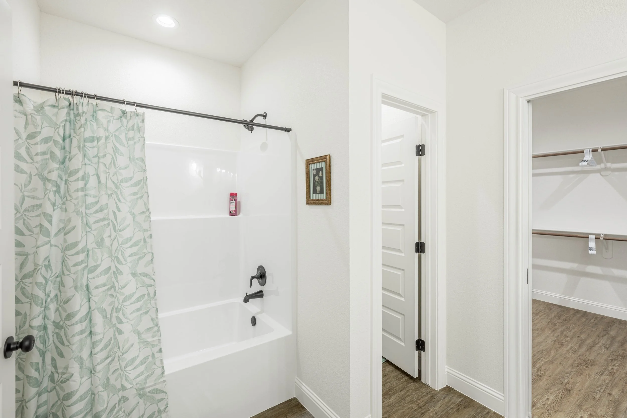 Bathroom with a white bathtub, shower curtain with leaf pattern, small framed artwork on the wall, and an open closet with a hanging rod and hangers.