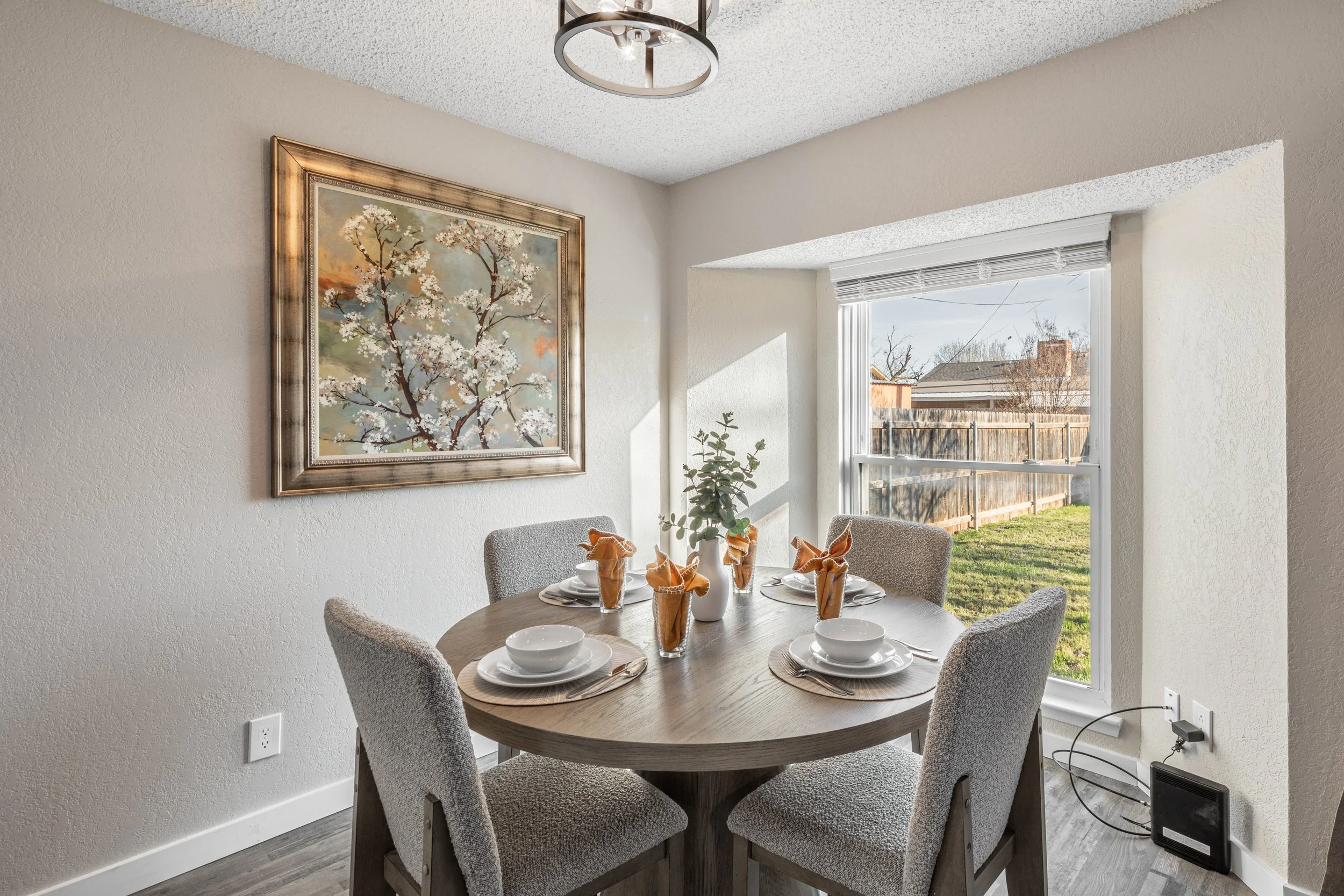 A dining area with a round wooden table, six upholstered gray chairs, white place settings, folded orange napkins, a white pitcher, and a wall art painting of a flowering tree. Sunlight streams through a window overlooking a backyard with a wooden fe