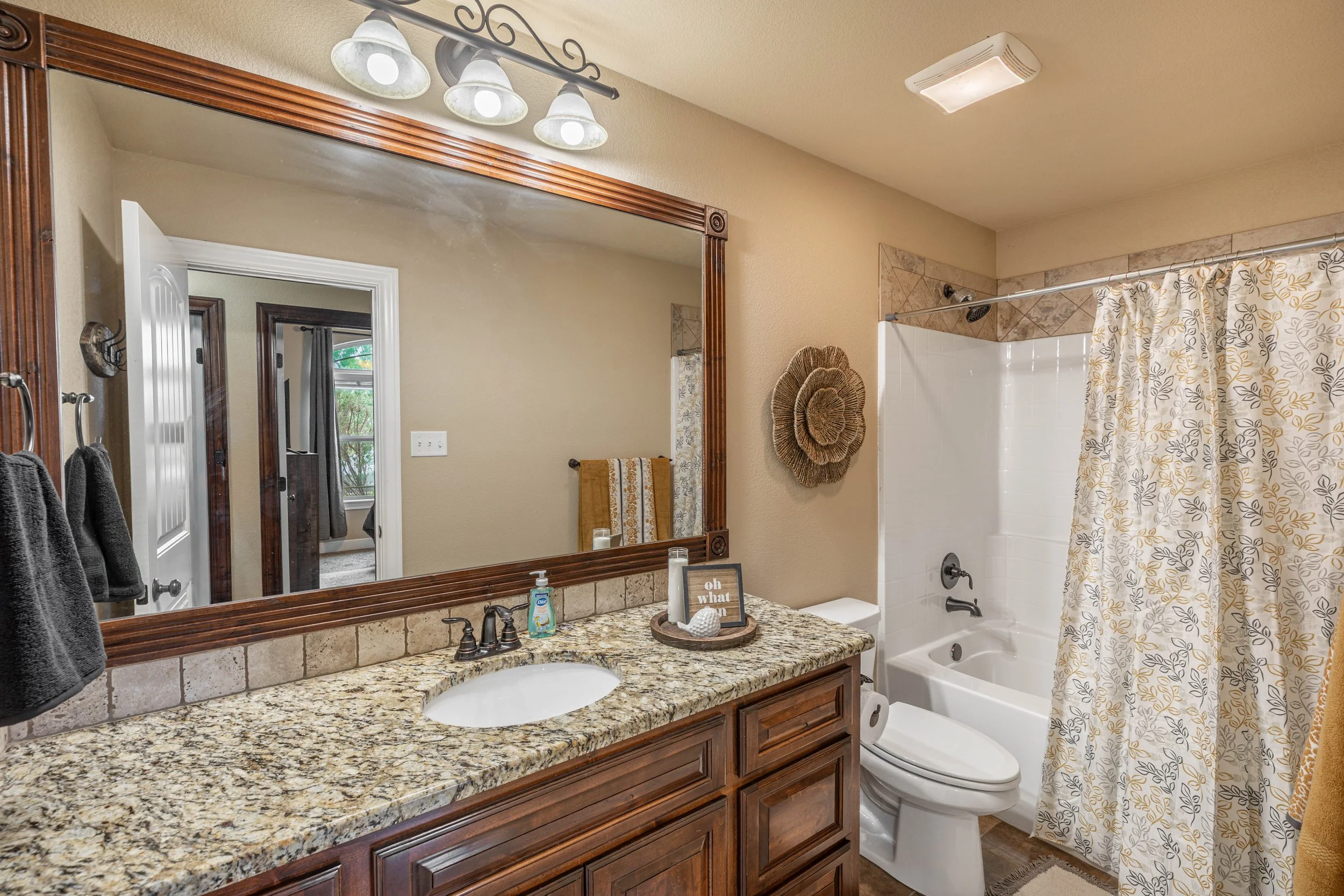 Bathroom with a granite countertop vanity, large mirror, three-light fixture, and a white bathtub with a curtain featuring yellow and gray leaf patterns. There are towels on hooks and decorative items on the counter, and a thermostat on the wall.