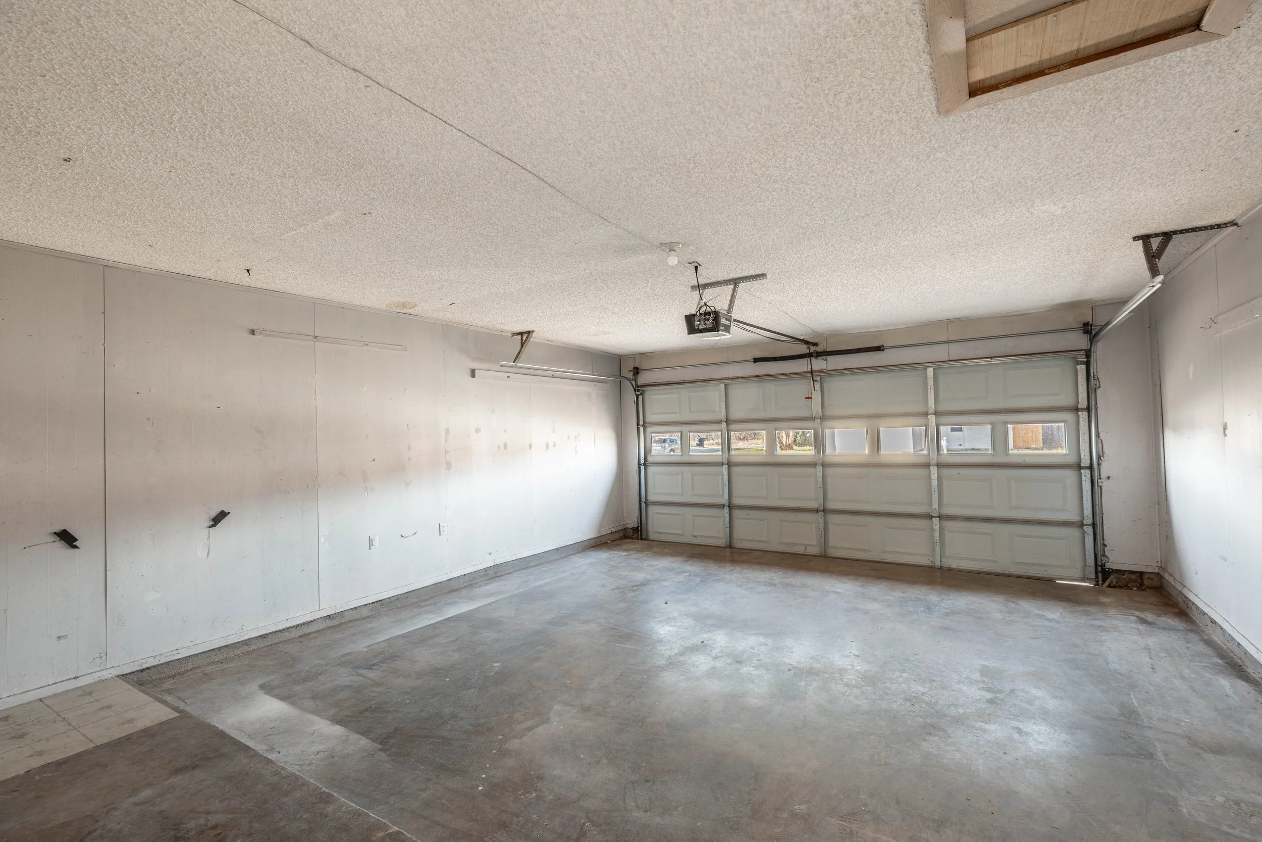 Empty garage with a large white garage door, concrete floor, and unfinished ceiling with a garage door opener and electrical wiring visible.