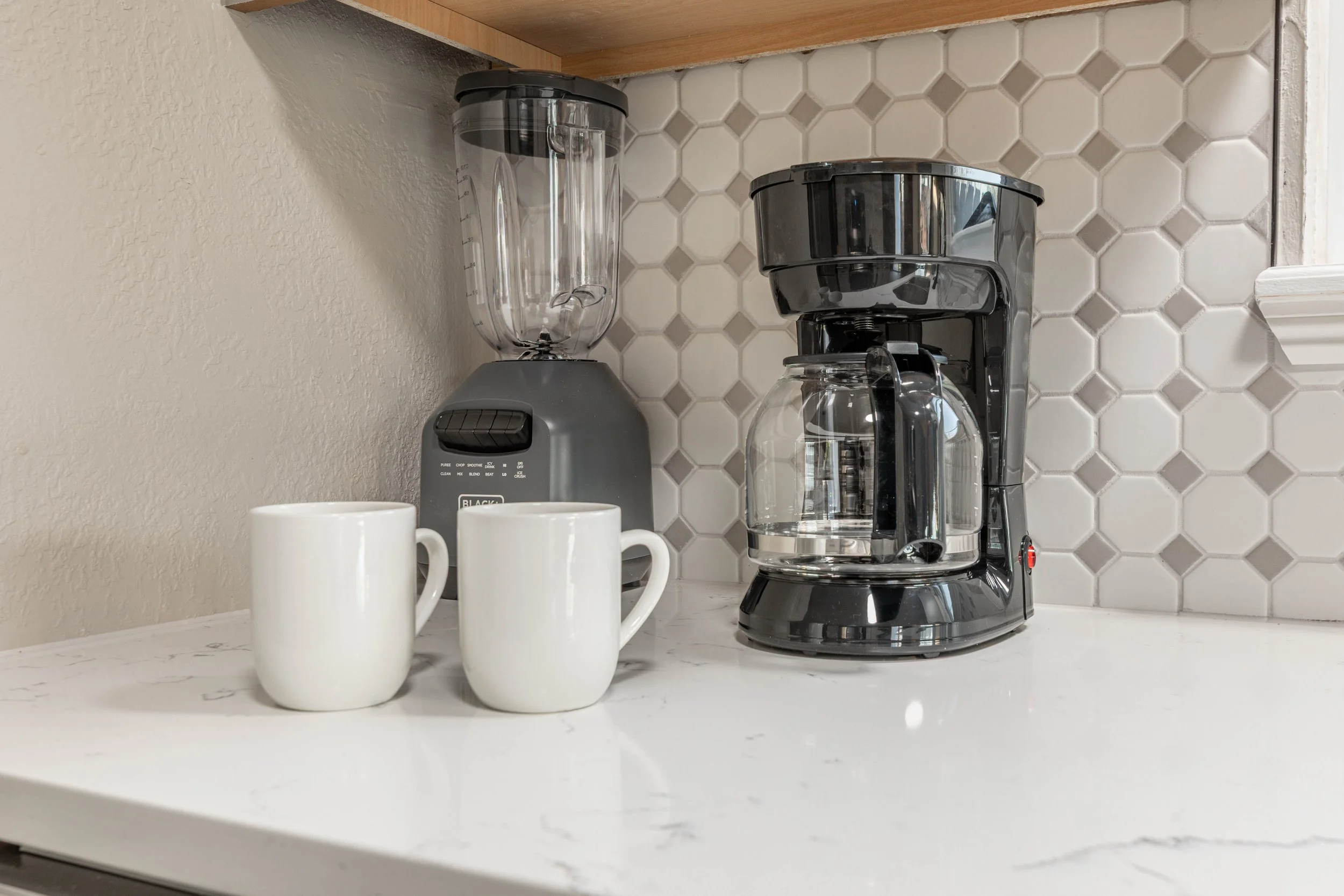 Two white coffee mugs, a black coffee maker, and a blender on a white kitchen countertop with a tiled backsplash.