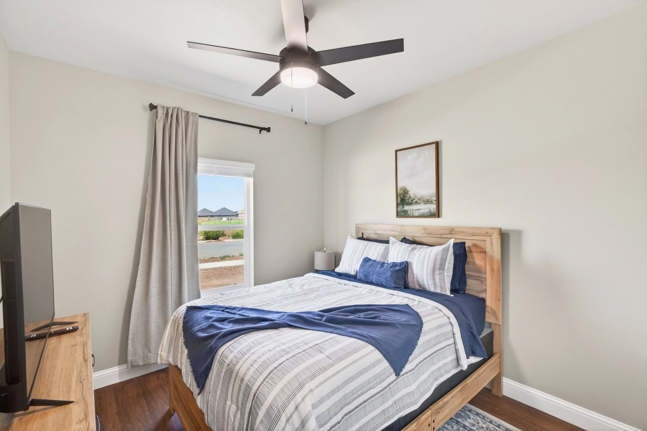 A bedroom with a wooden bed, white and blue striped bedding, a nightstand with a lamp, a window with beige curtains, a framed landscape picture on the wall, a ceiling fan with lights, and a partial view of a TV.
