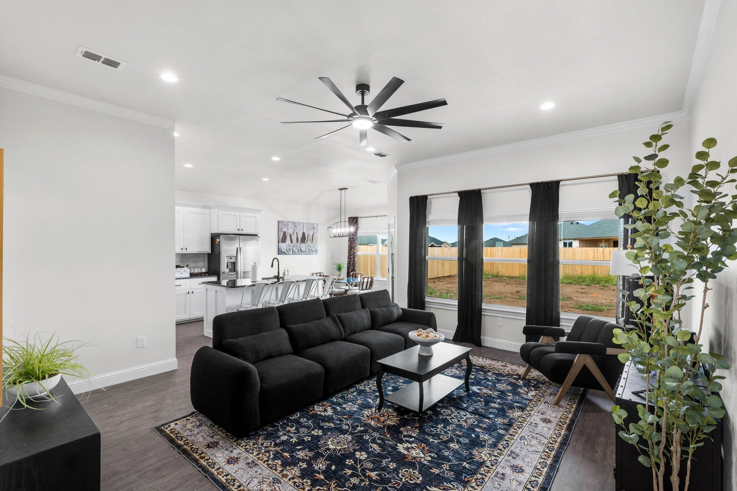 Living room with large black sofa, coffee table, and decorative rug, open to kitchen, large windows with black curtains, ceiling fan, and potted plant.