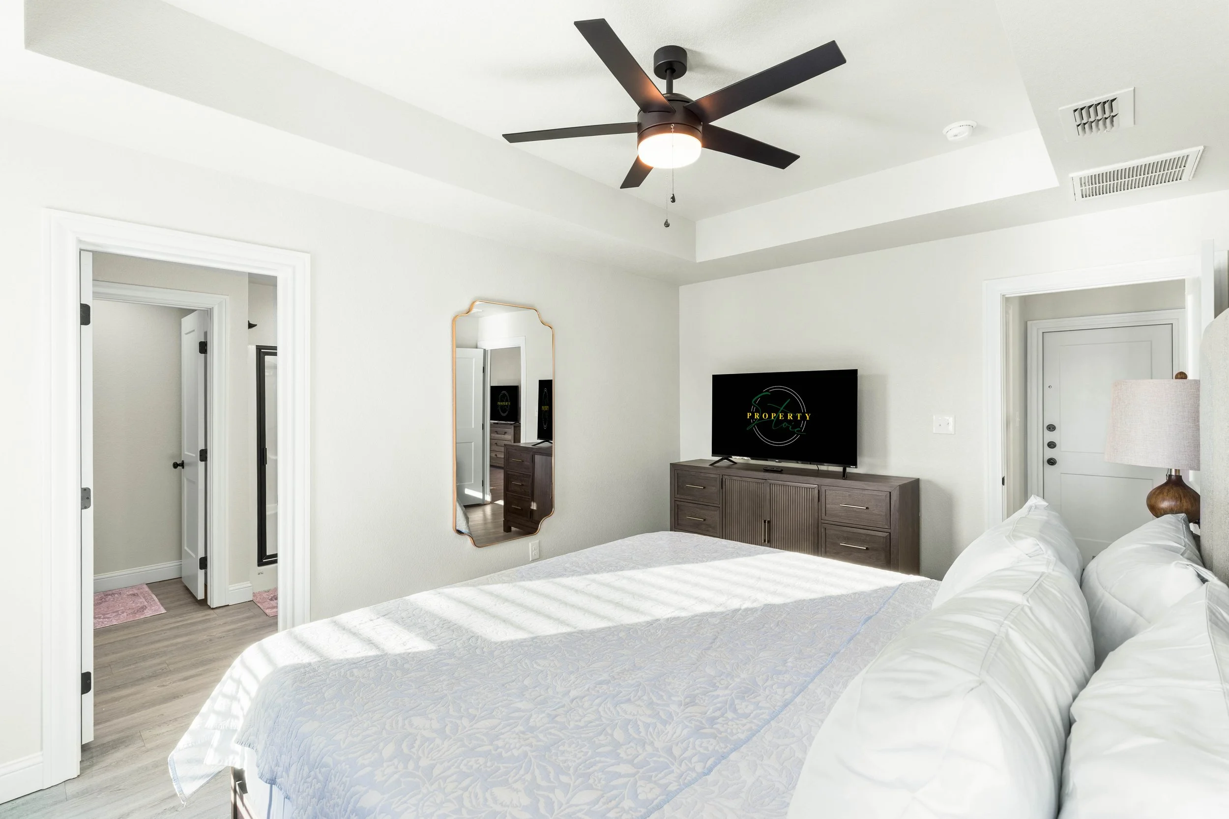 A bedroom with a bed, a dresser with a TV on top, a wall mirror, a ceiling fan, and a doorway. The room has white walls and a light wood floor.