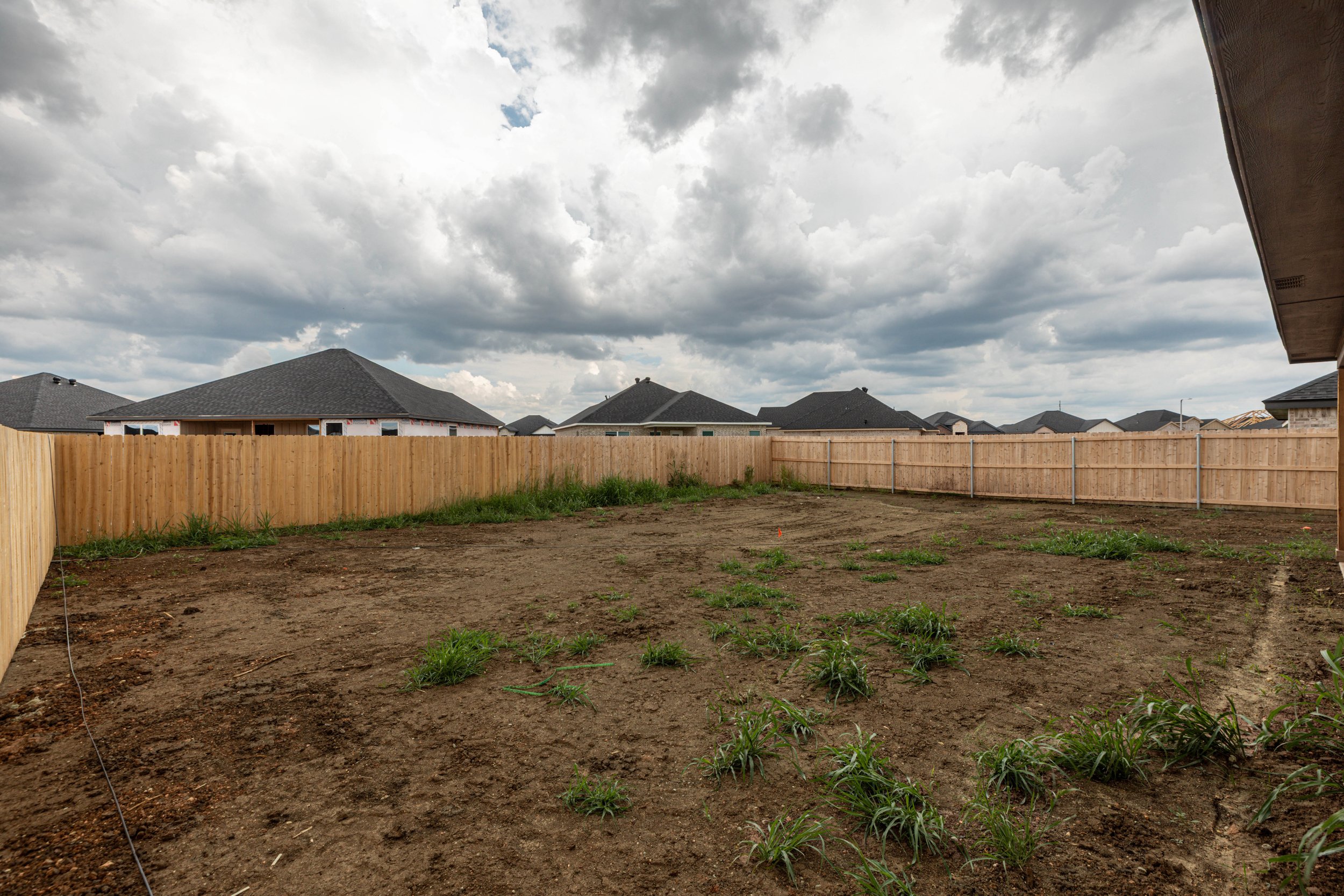 View of a backyard with a dirt patch and small grass patches, enclosed by a wooden fence, with new houses with dark roofs in the distance under a cloudy sky.