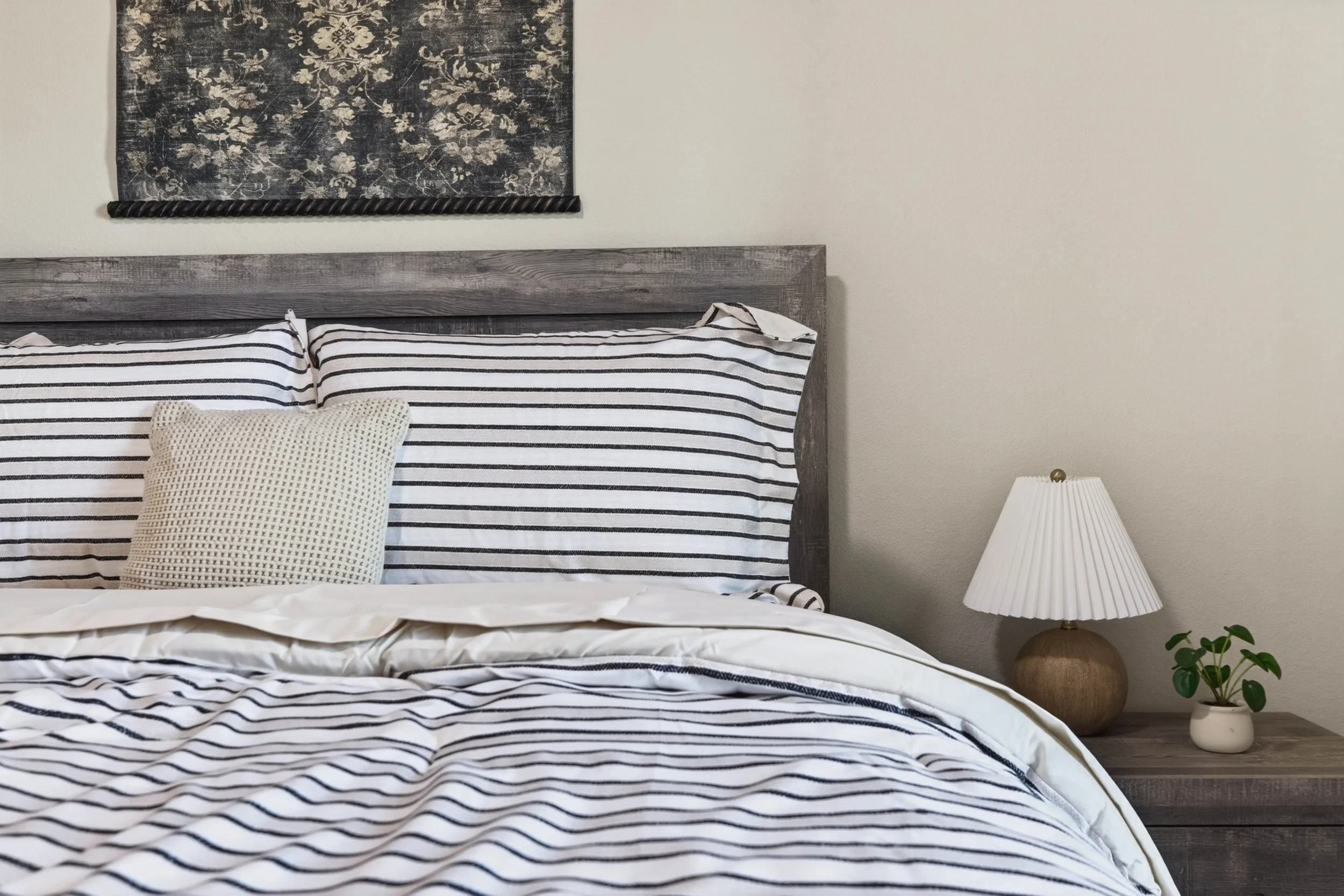 A neatly made bed with black and white striped sheets, a tan textured pillow, and a wooden headboard. To the right, a white table lamp with a pleated shade sits on a small wooden nightstand, beside a potted green plant.