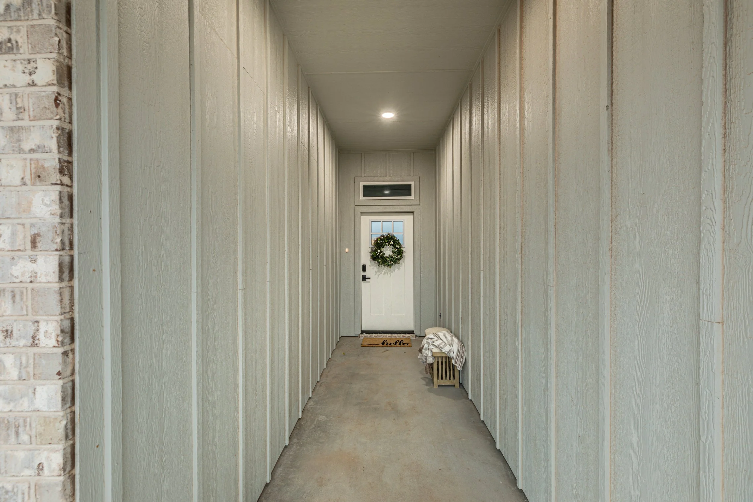A long hallway painted light green with vertical paneling, leading to a white door adorned with a holiday wreath. The hallway has a small bench and a 'hello' welcome mat at the entry.
