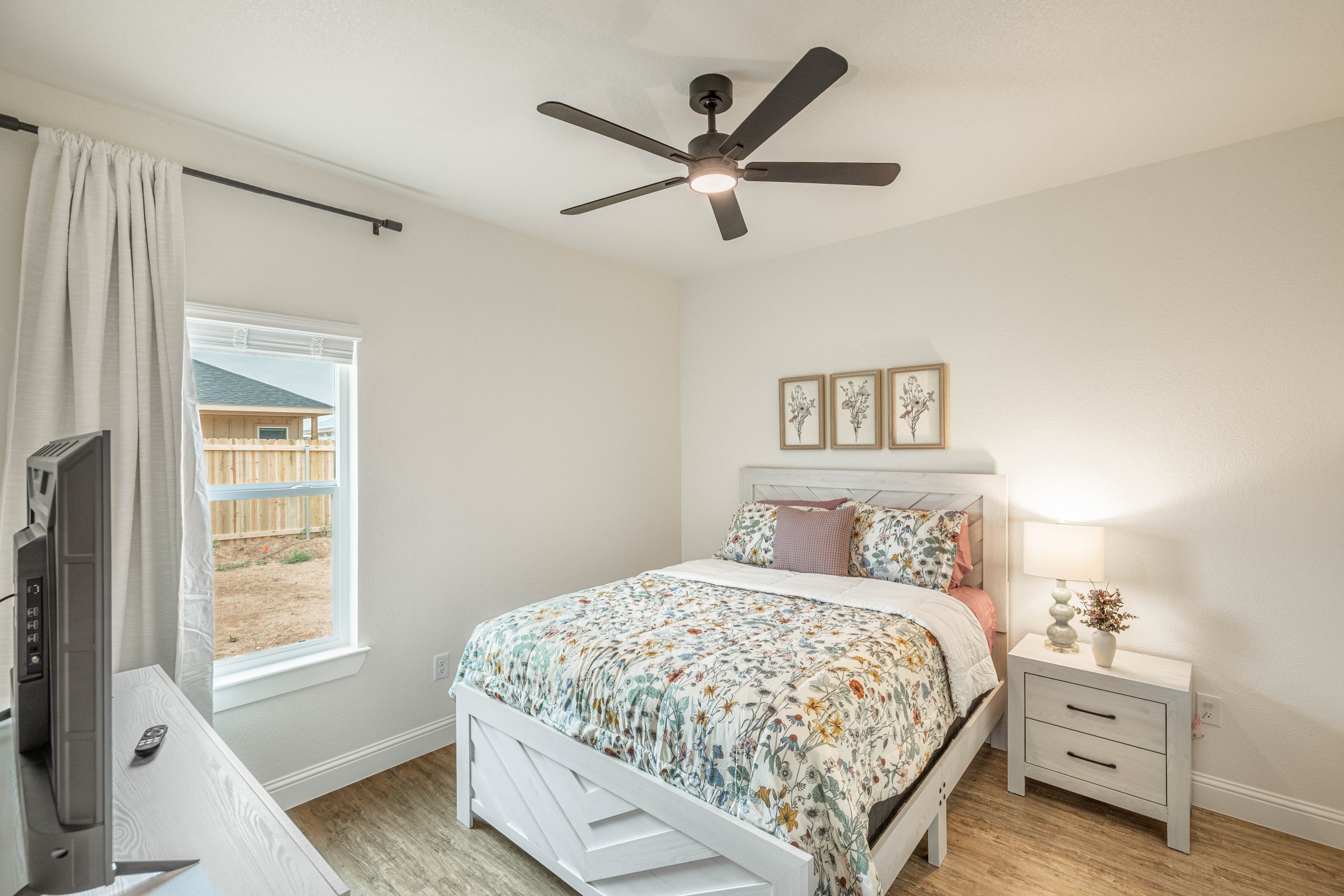 Bedroom with a white bed, floral bedspread, framed botanical art above the bed, white nightstand with a lamp, window with white curtains, ceiling fan with light, and a wooden fence outside.