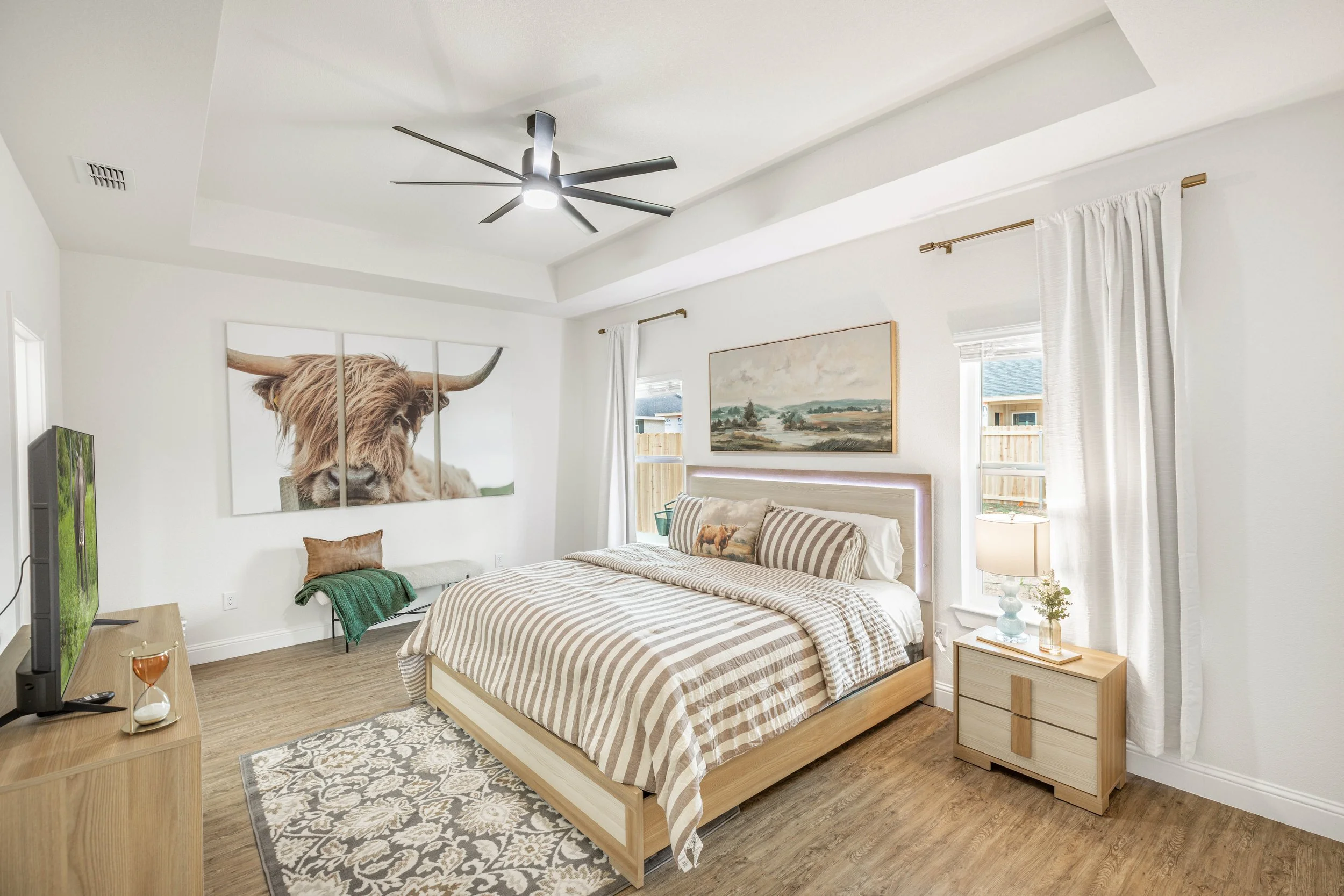 A bedroom with a large bed, striped bedding, and pillows, a nightstand with a lamp, and artwork of a highland cow and landscape on the walls, with windows and curtains, and a ceiling fan.