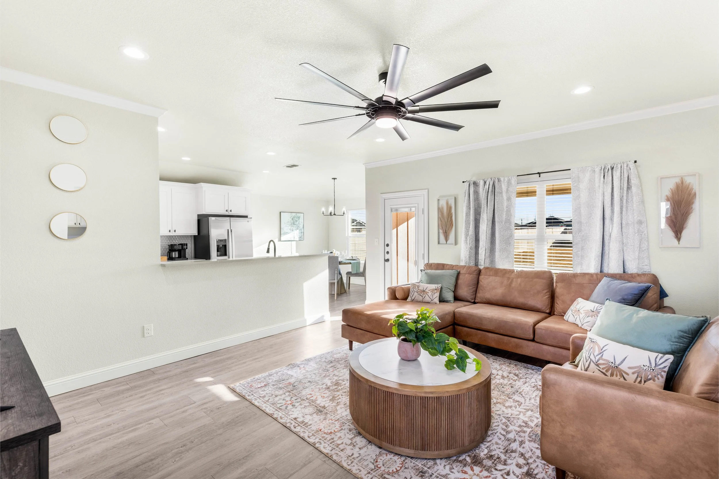 Living room with beige sofa, round coffee table, houseplant, large windows with curtains, kitchen in background, ceiling fan, wall art, and wooden flooring.