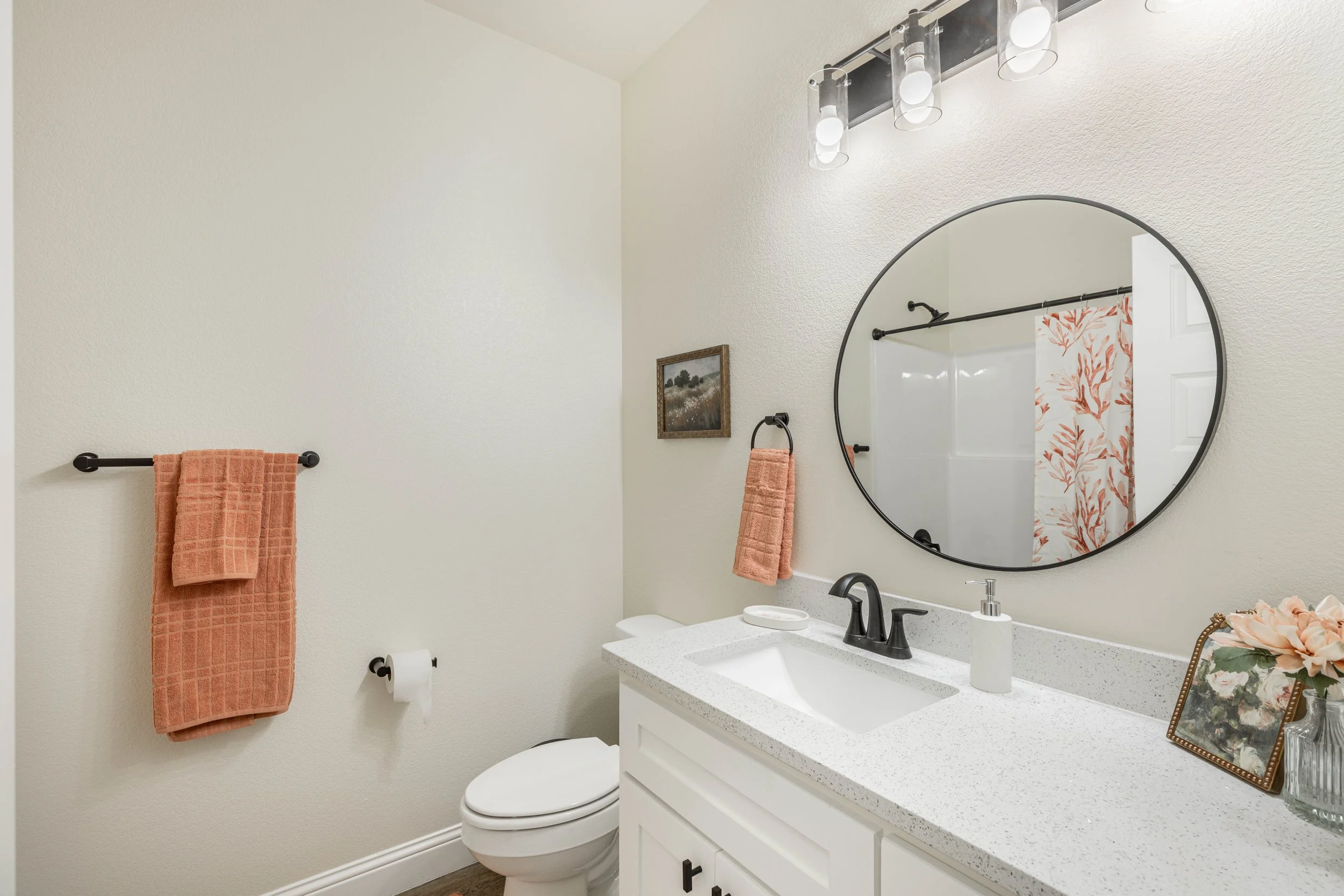 A clean bathroom with a white vanity, black fixtures, a round mirror, pink towels on a black towel bar, a framed landscape picture, a tissue box, and a vase with flowers on the counter. There is a shower curtain with coral-colored leaves visible in t