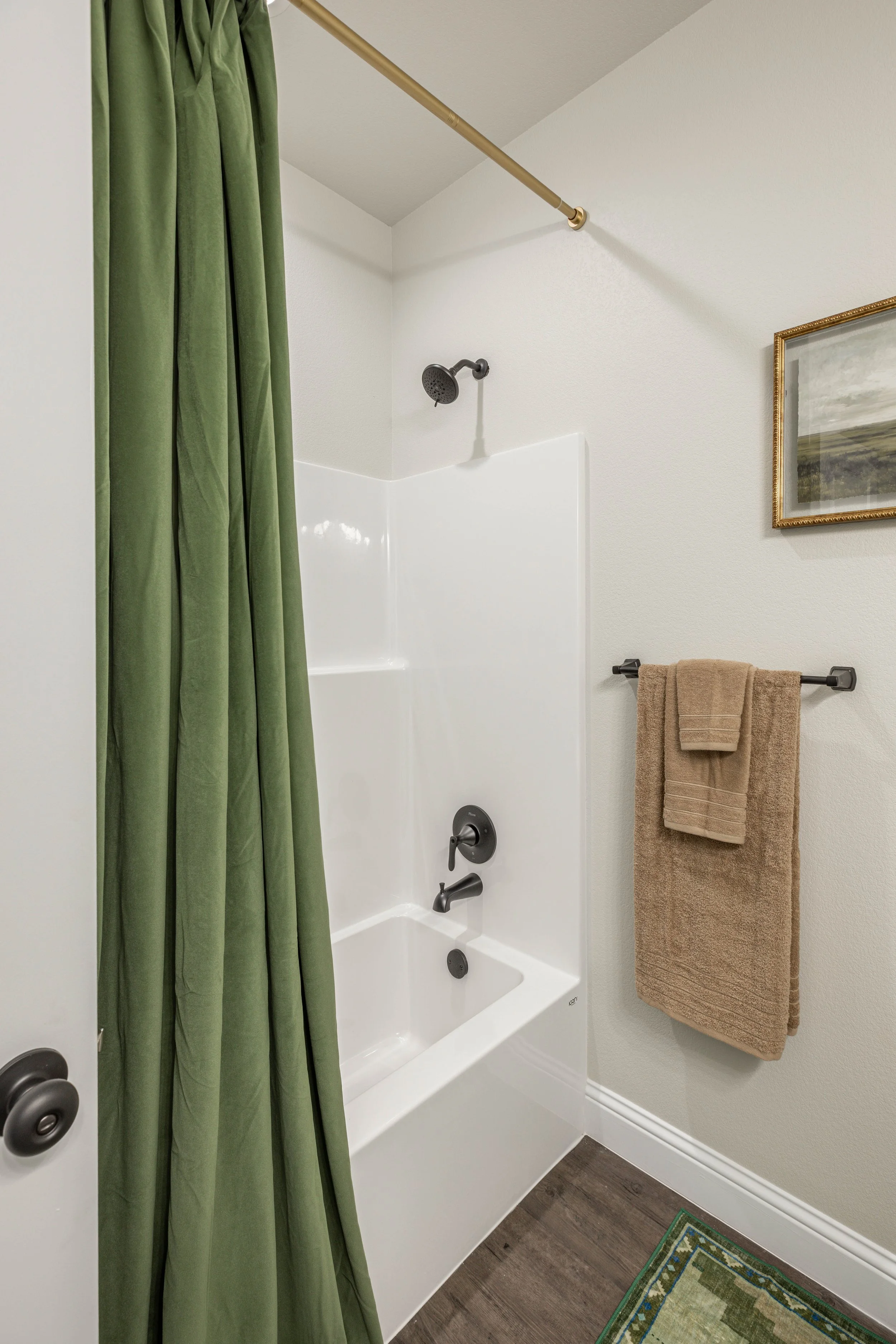 Bathroom with a white bathtub and shower, a green shower curtain on a curved rod, tan towels on a black towel rack, a framed landscape picture on the wall, dark wood flooring, and a patterned rug.