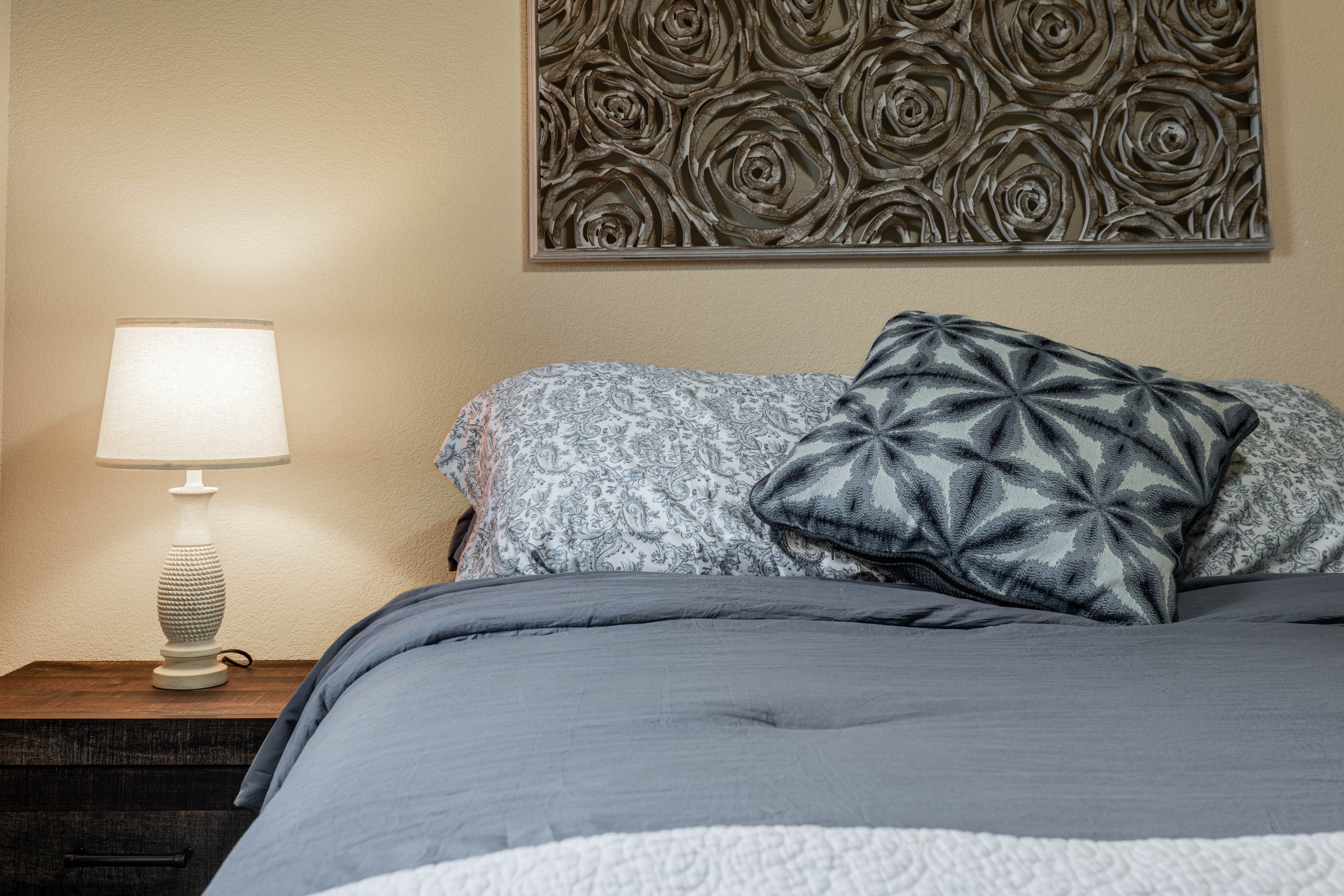 A bedroom with beige walls, a nightstand, and a table lamp. There are patterned pillows on the bed, including one with a dark floral design, and a neatly made bed with a blue and white comforter. A decorative wall art piece with a floral motif is hun