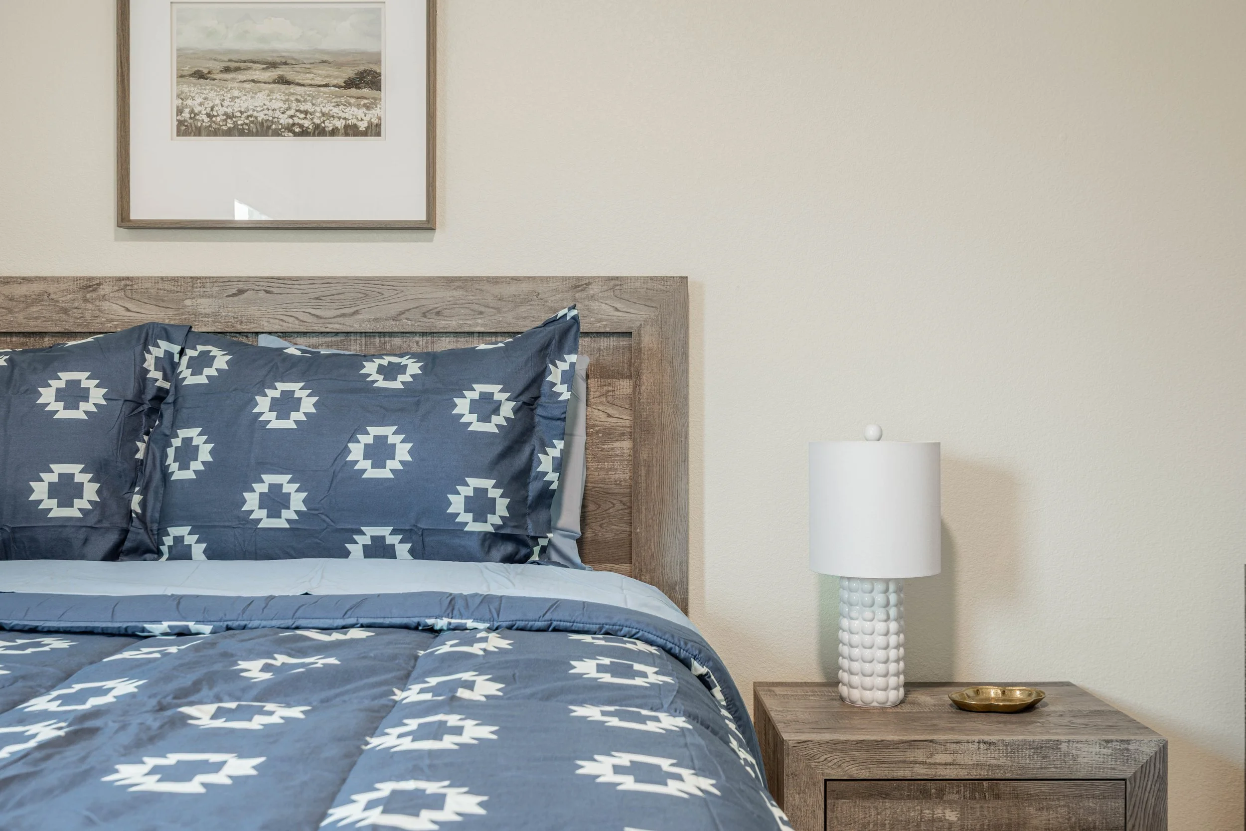 A bedroom scene featuring a bed with blue and white patterned bedding, a wooden headboard, a minimalist white lamp on a wooden nightstand with a small gold tray, a neutral-colored wall with a landscape painting, and a plain beige wall.