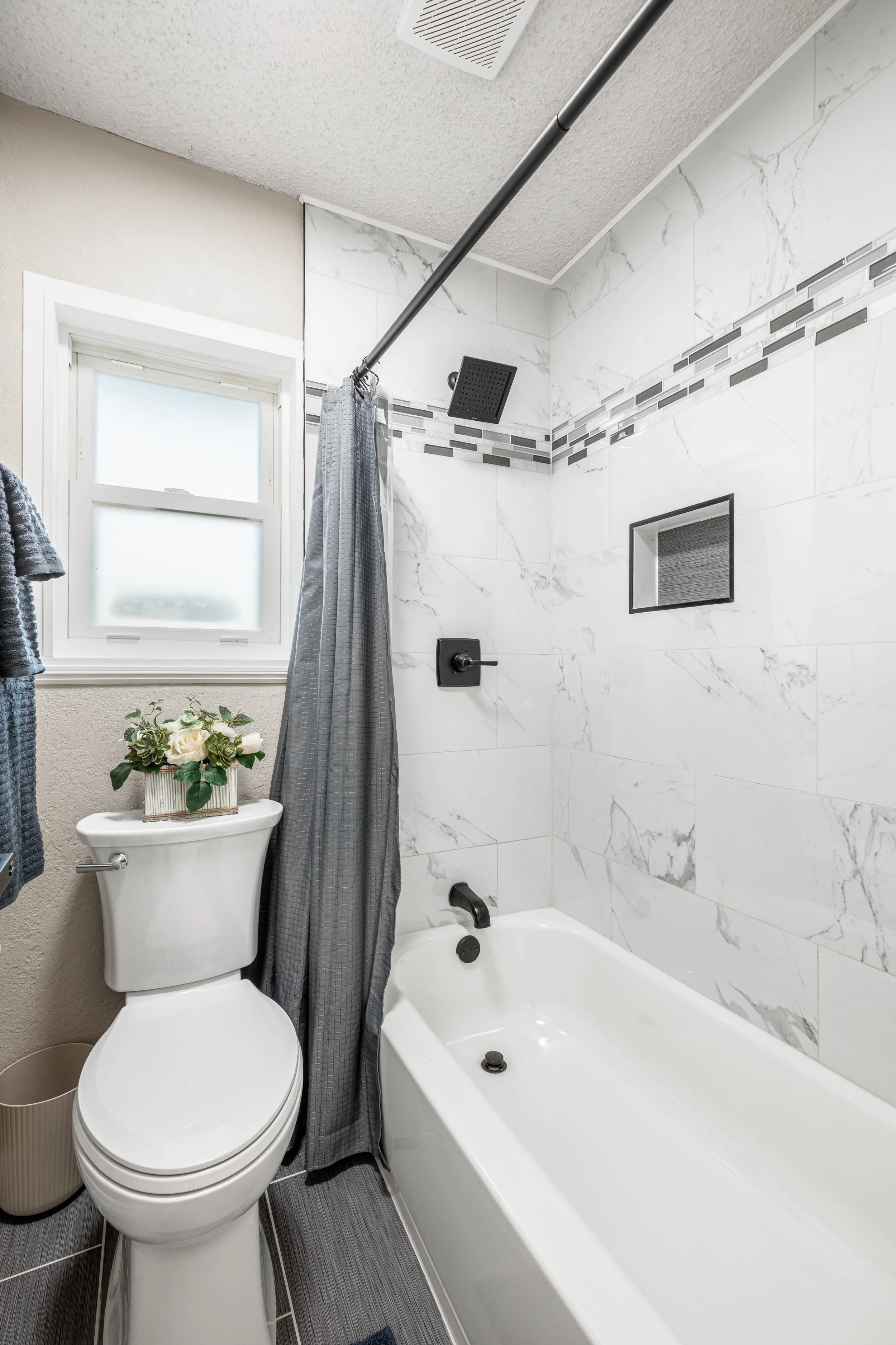 A modern bathroom with a toilet, a bathtub, and a shower area. The shower has a square black rain shower head, a gray curtain, and a tiling wall with marble-like design and decorative strip. There is a window with frosted glass and a flower arrangeme