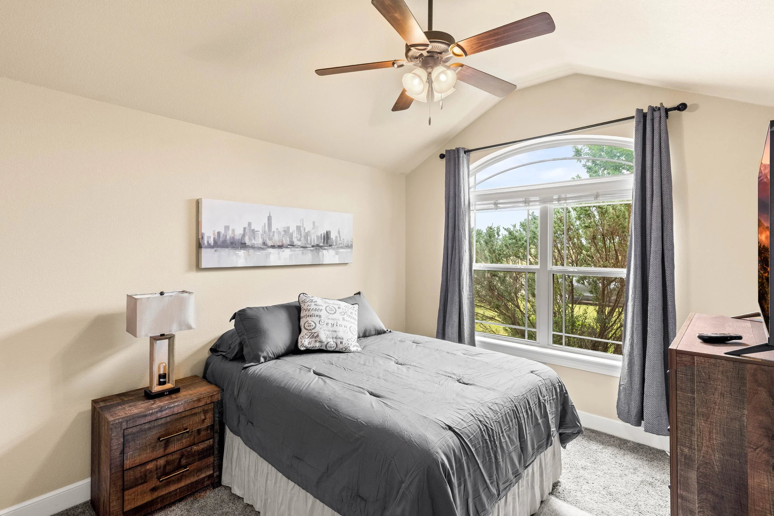 A bedroom with beige walls, a large arched window with blue curtains, a bed with dark gray bedding, a white pillow with writing, wooden nightstands, a table lamp, a ceiling fan with lights, and outdoor greenery visible through the window.