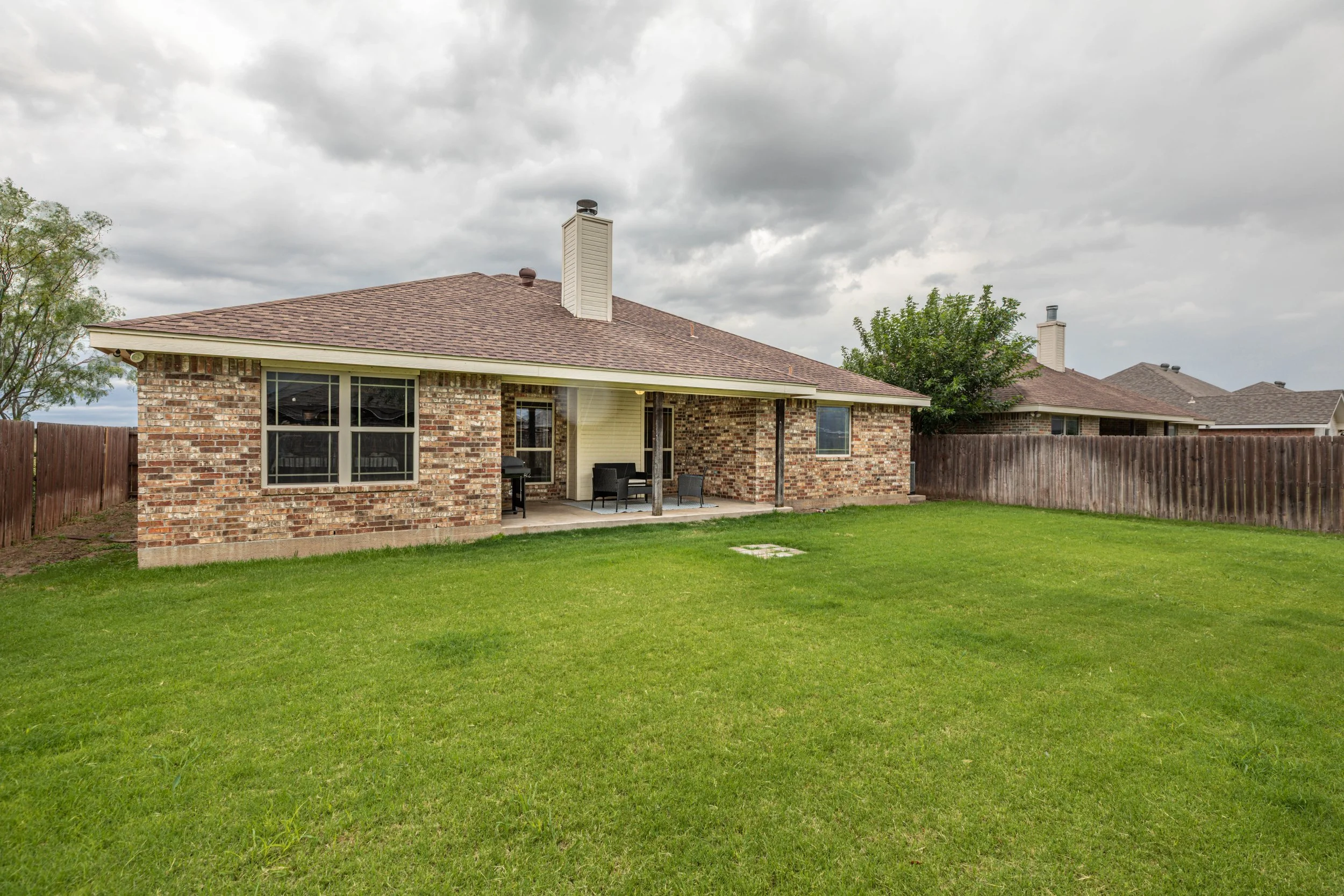 Backyard view of a brick house with patio furniture, surrounded by a wooden fence and green grass, under cloudy sky.