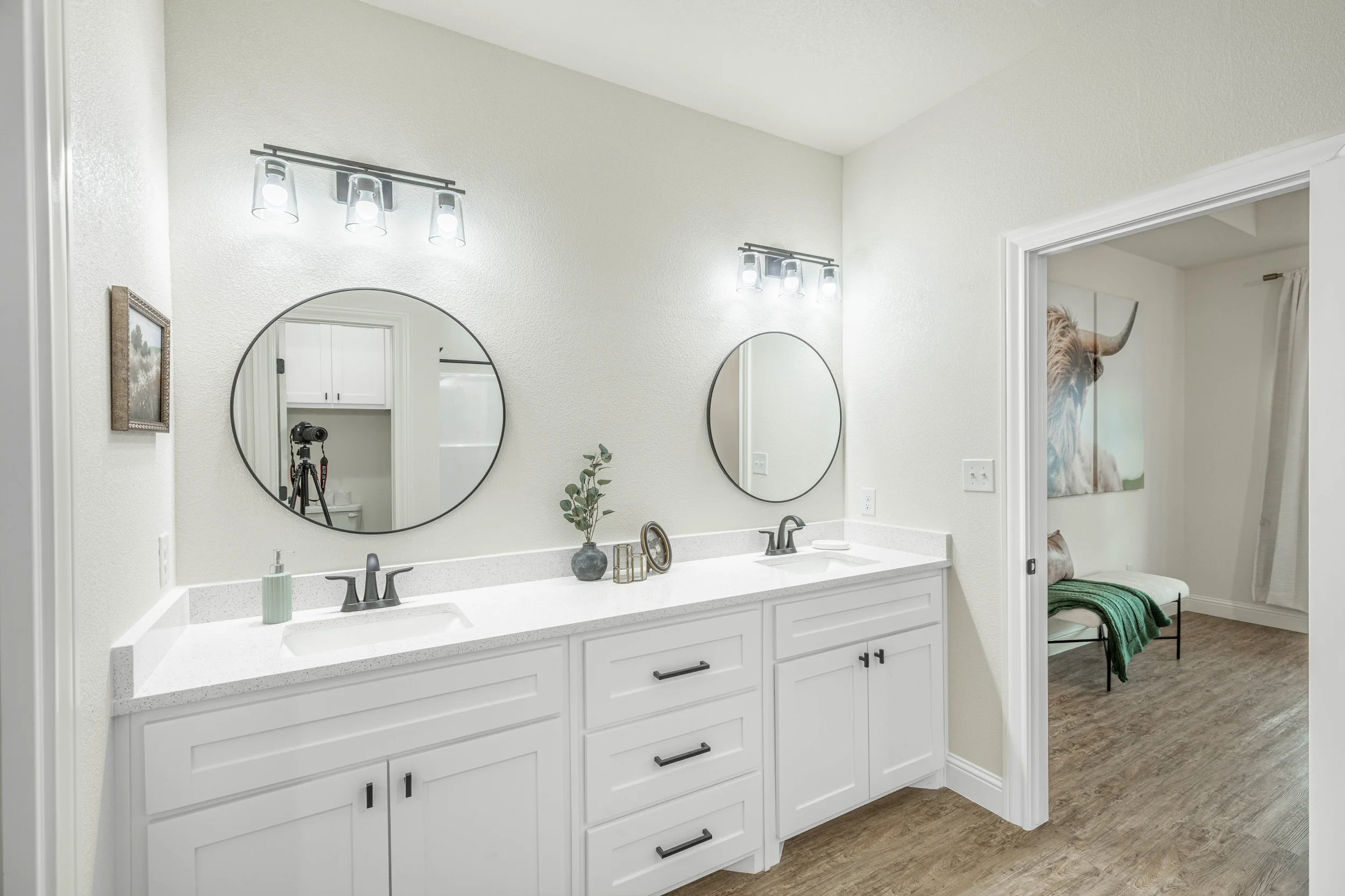 A modern bathroom double vanity with two round mirrors, a white countertop, two sinks, and contemporary black hardware. Decor includes a small plant, candle, and decorative items. Overhead lighting fixtures with multiple bulbs illuminate the space. A