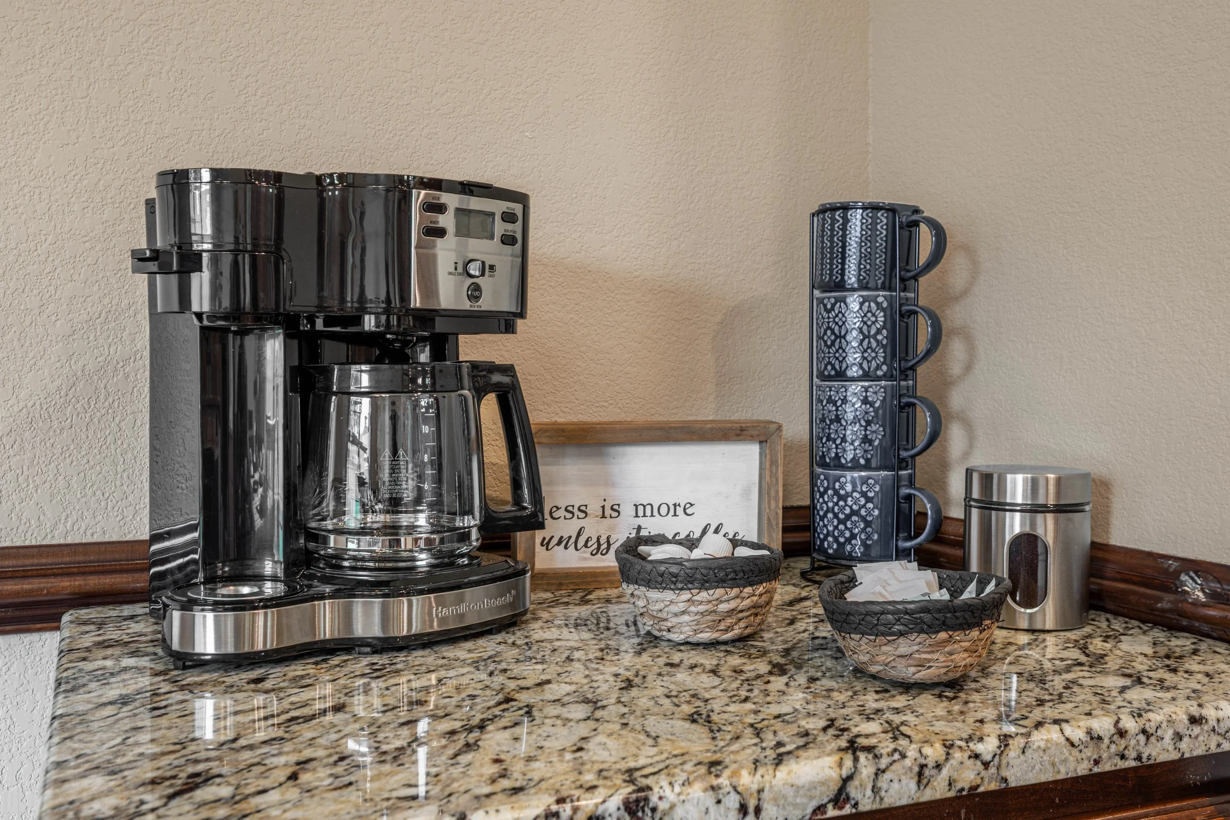 A coffee maker on a speckled granite kitchen counter next to black and white patterned mugs, a stainless steel container, and decorative baskets, with a framed sign that reads, 'Less is more unless it’s coffee.'