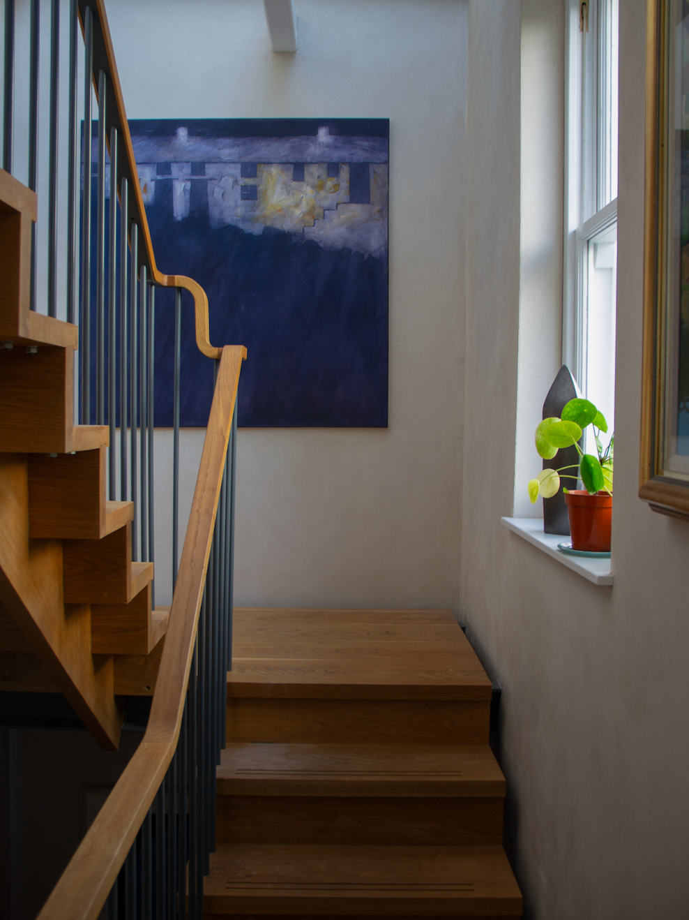 Wooden staircase with black railing, a blue abstract painting on a white wall, and a window with a potted plant on the sill.