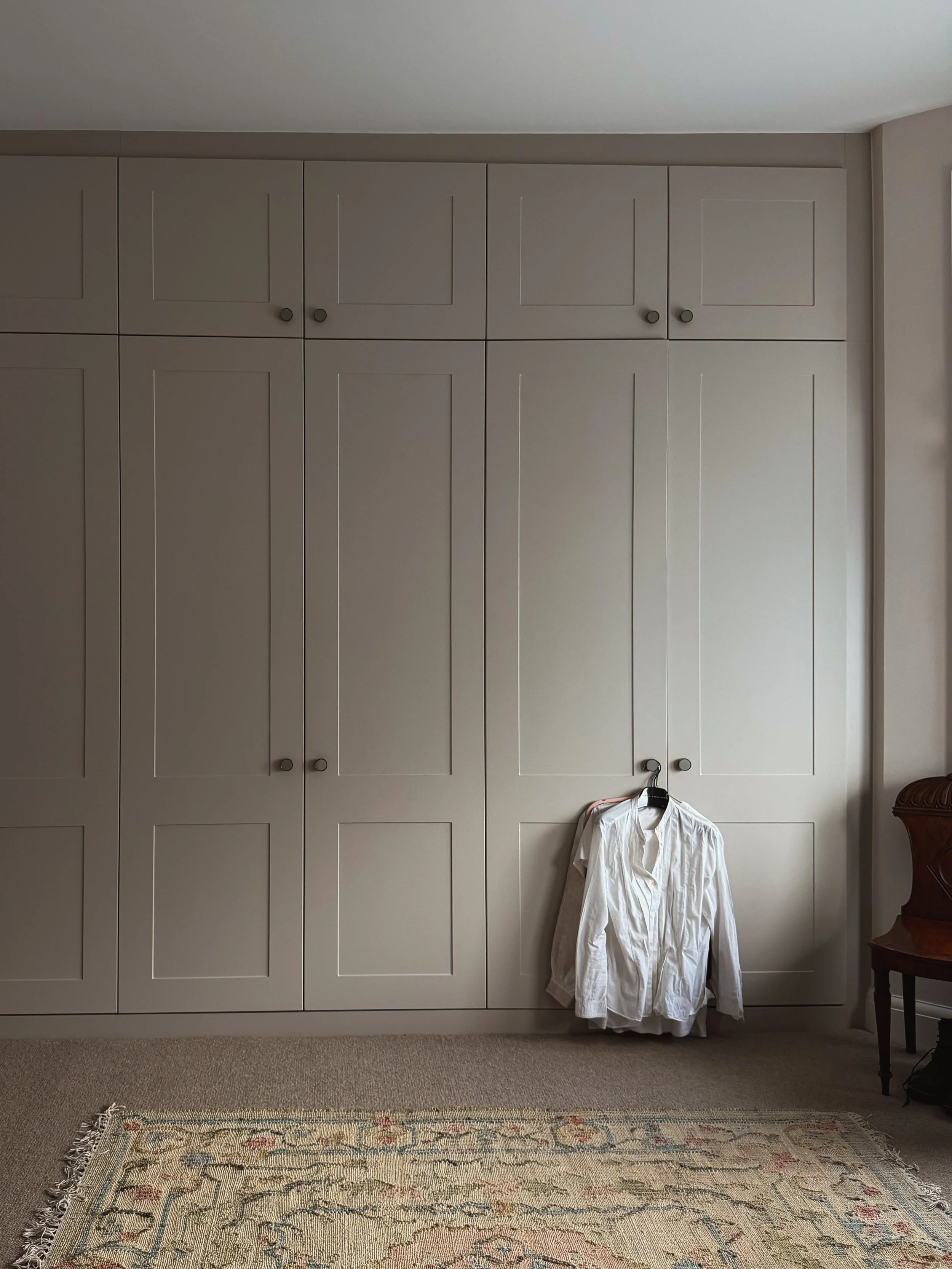 A white dress shirt hanging over a chair in front of a beige built-in wardrobe with closed doors, in a room with a wooden side table, a patterned rug, and light-colored walls.