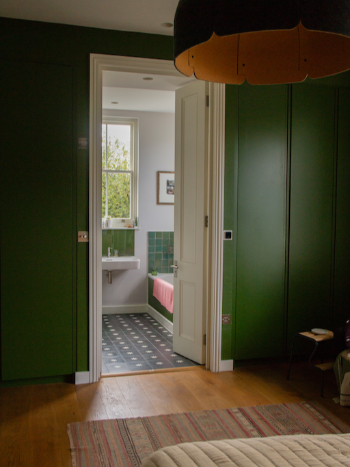 A doorway leading to a bathroom with a window, sink, and bathtub with a pink cover, green tiles, and patterned black and white floor, visible through the open door in a room with dark green walls and wooden floor.