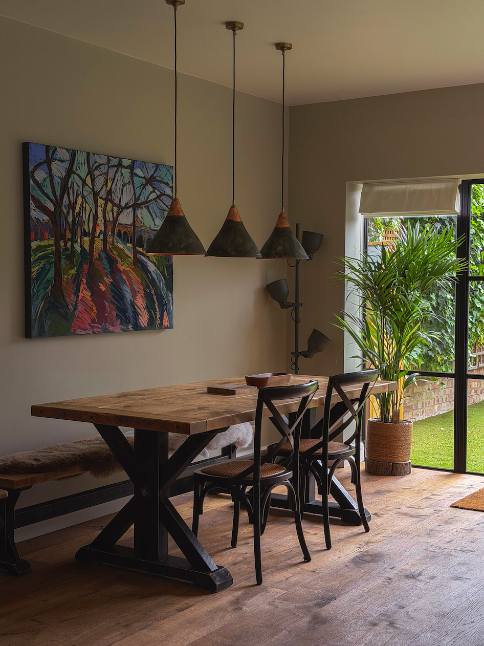 A dining area with a wooden table and three black chairs. There are three black pendant lights hanging above the table, a painting on the wall, a potted plant near a large window, and a natural wood floor.
