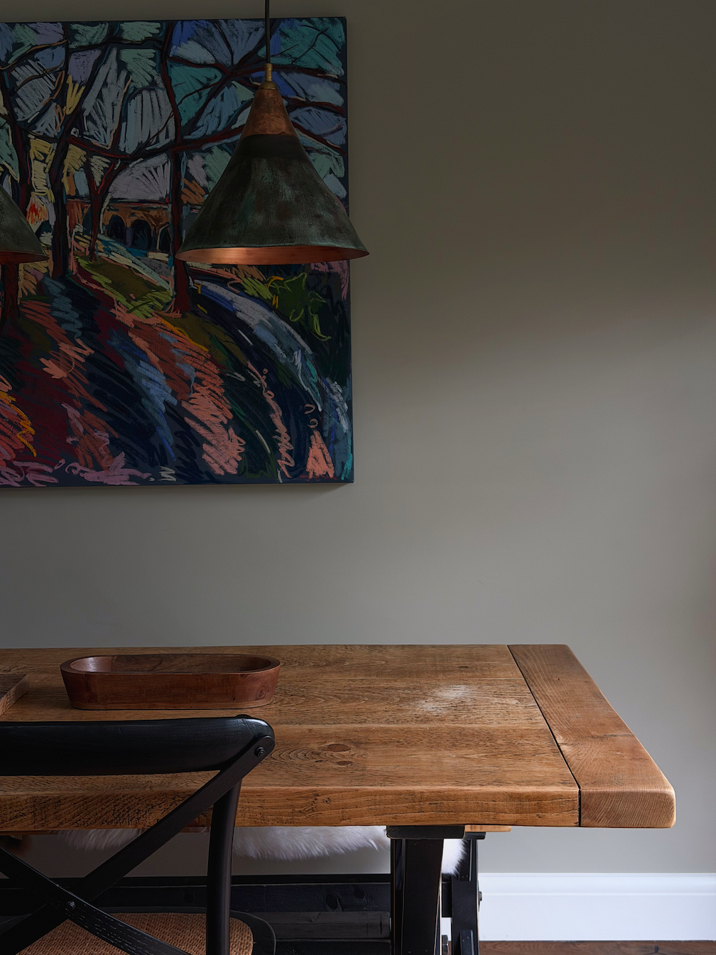 A wooden dining table with a black chair in front of it. Two industrial-style pendant lights hang above the table, and an abstract colorful painting is on the wall behind.