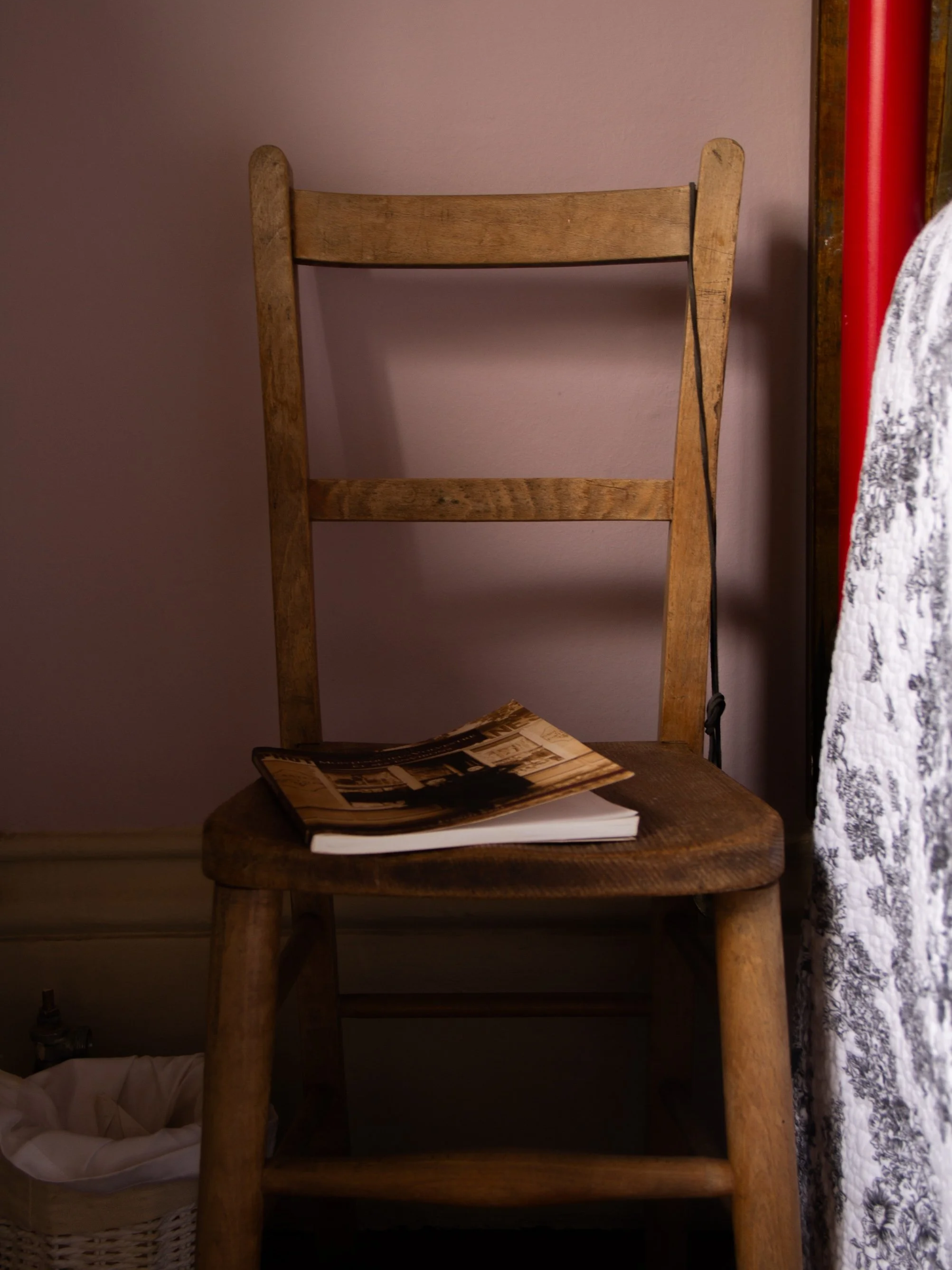 A wooden chair with a book and magazine on its seat, placed in front of a pinkish wall, with a basket and a patterned cloth visible to the sides.