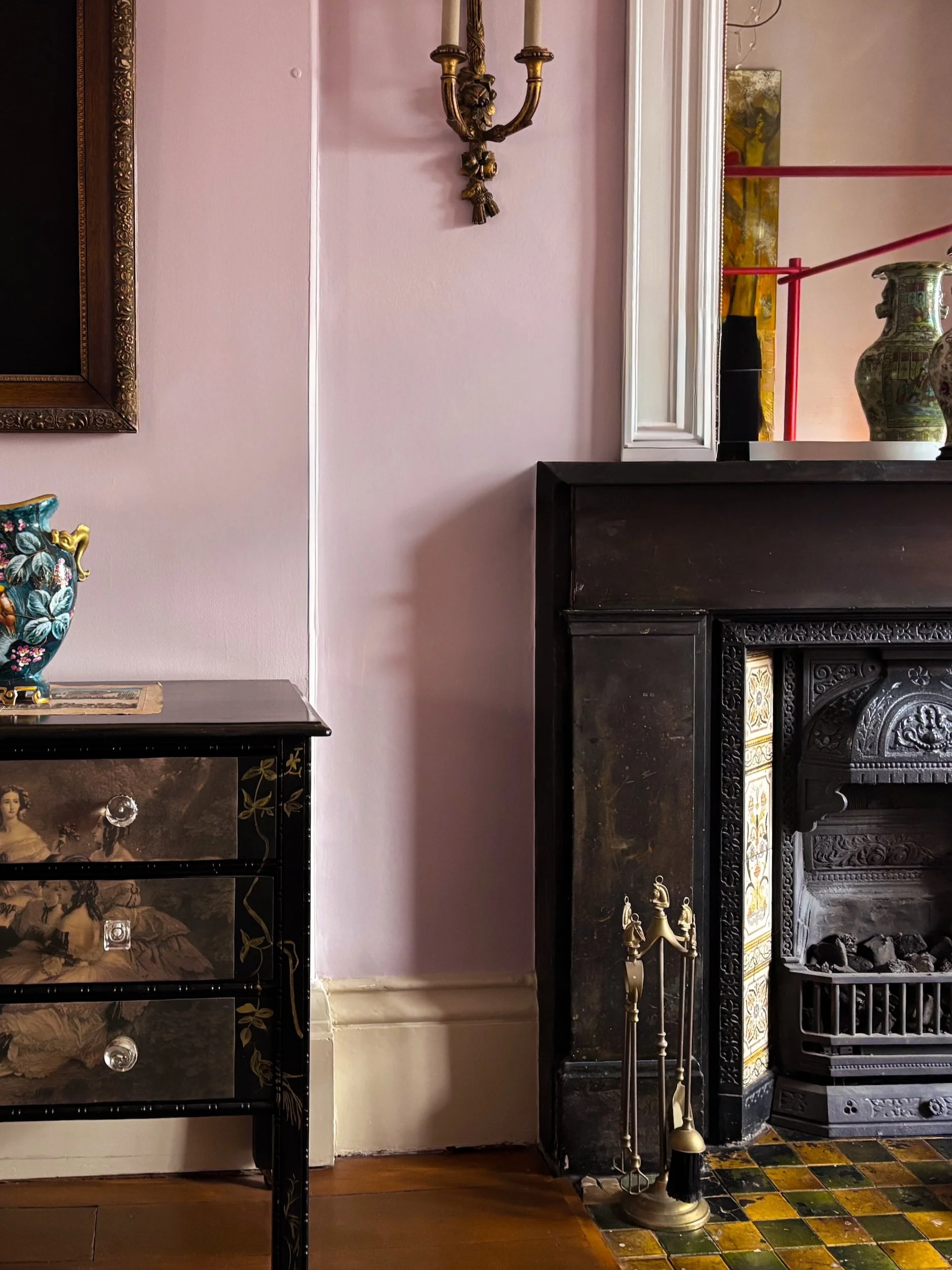Interior of a room with a pink wall, a black fireplace with ornate details, a tall candlestick with brass candlesticks, objects on top of a black chest of drawers, and a colorful tiled floor.