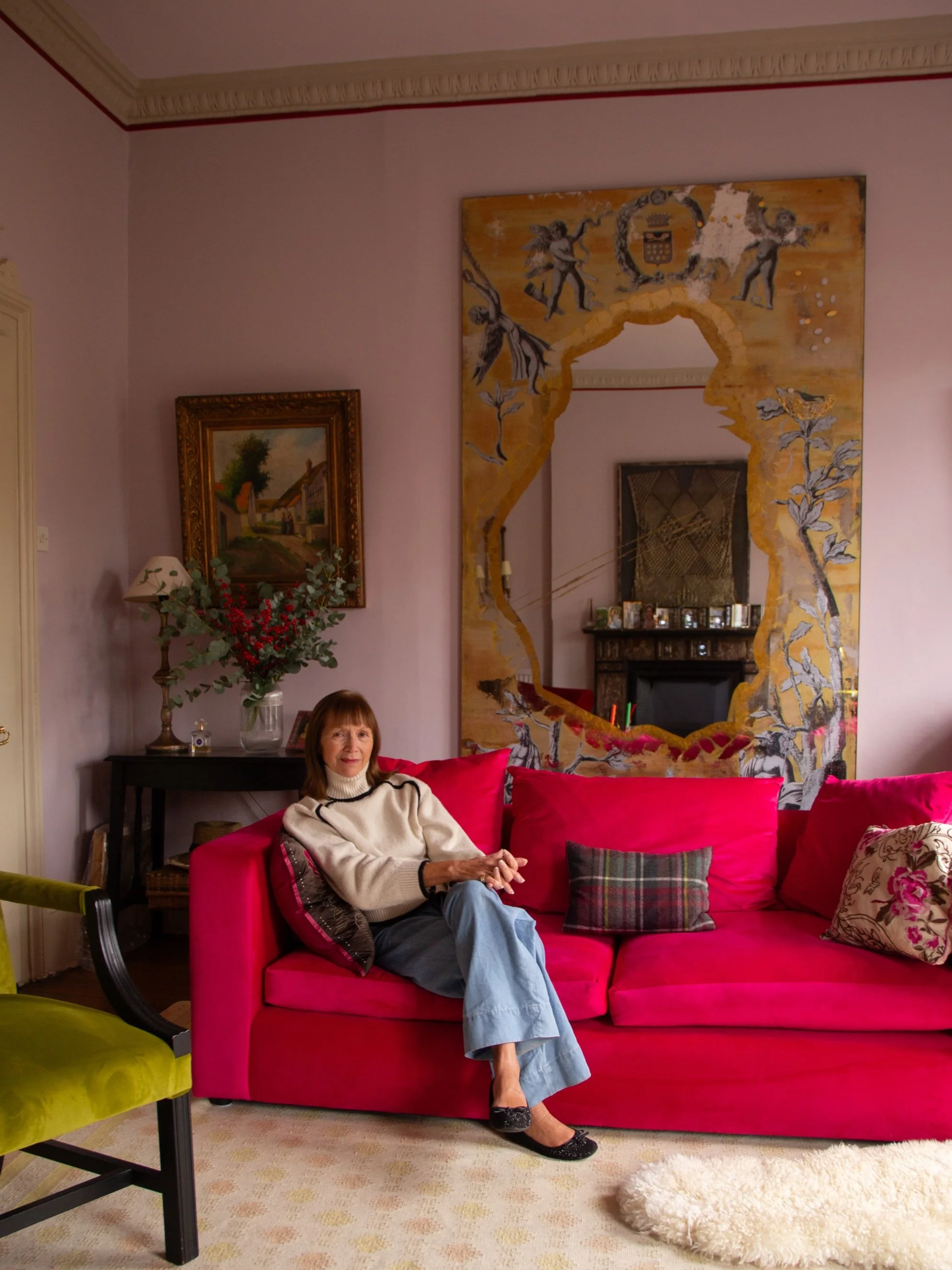 A middle-aged woman with brown hair and bangs, wearing a cream sweater with black stripes, light blue jeans, and black shoes, is sitting on a bright pink sofa in a living room. She is smiling with her hands clasped on her lap, and the room has a beige carpet, a dark wooden side table with a vase of red berries and a white lamp, a framed painting of a rural scene, and a decorative mirror with a gold frame with painted angels, a crest, and floral designs on the wall behind her.