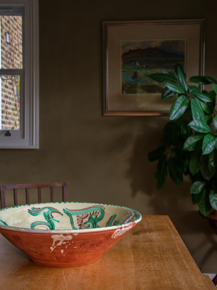 A ceramic bowl with colorful designs on a wooden table in a room with a window, a leafy plant, and framed artwork on the wall.