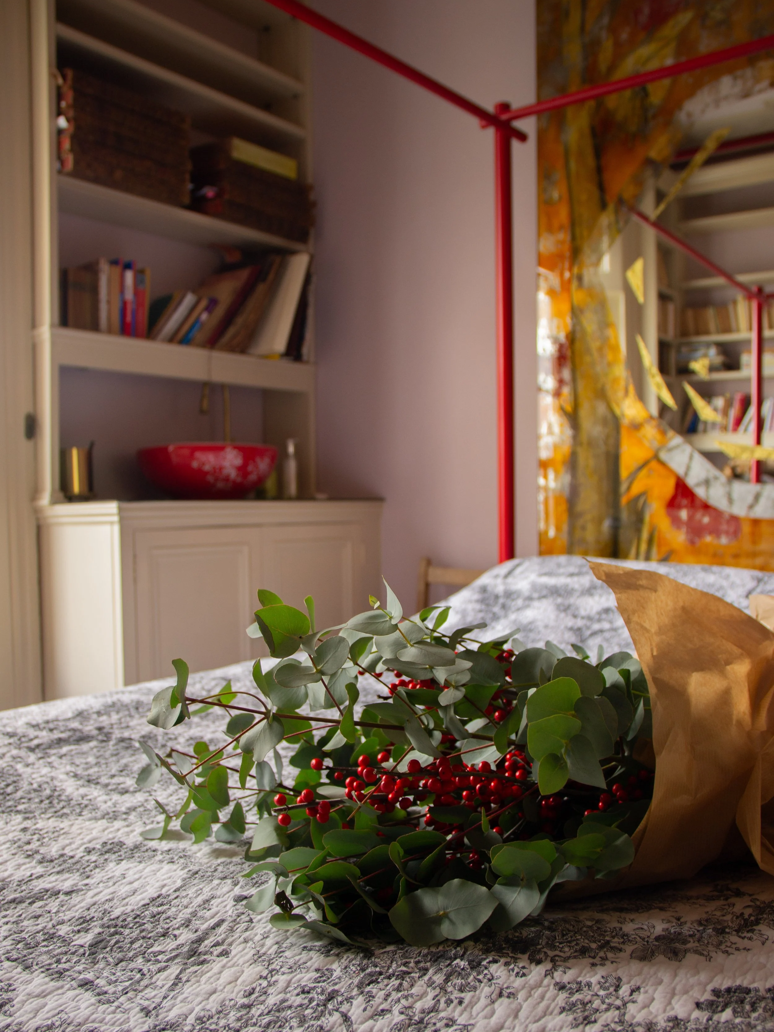 A close-up of a bouquet of greenery and red berries on a quilted bedspread with a floral pattern in a cozy bedroom. In the background, there are bookshelves with books and decorative items, and a window with yellow curtains.