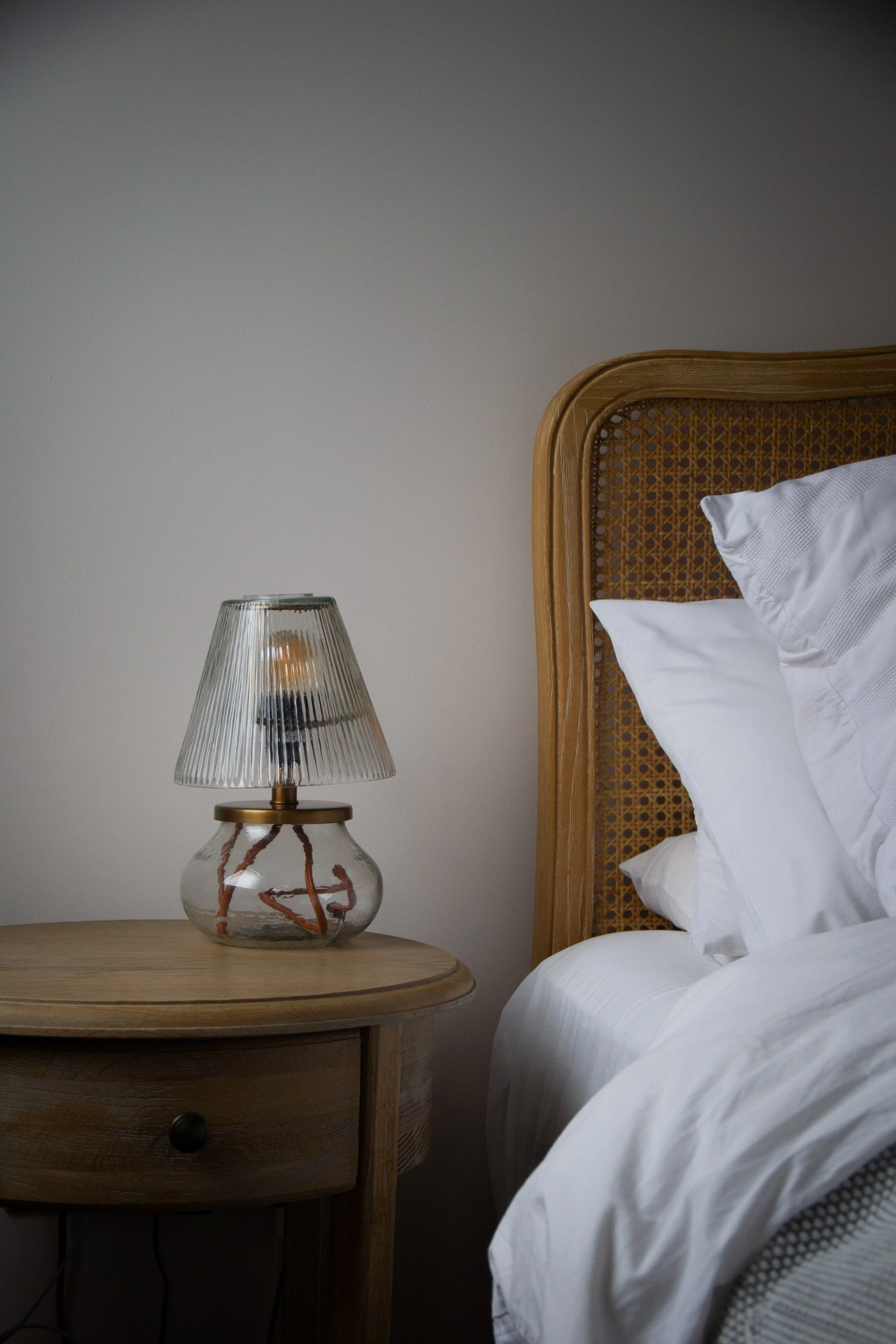 A bedroom corner with a wooden nightstand, a modern table lamp with a glass base and a ribbed glass shade, an unmade bed with white bedding, and a rattan headboard against a plain wall.