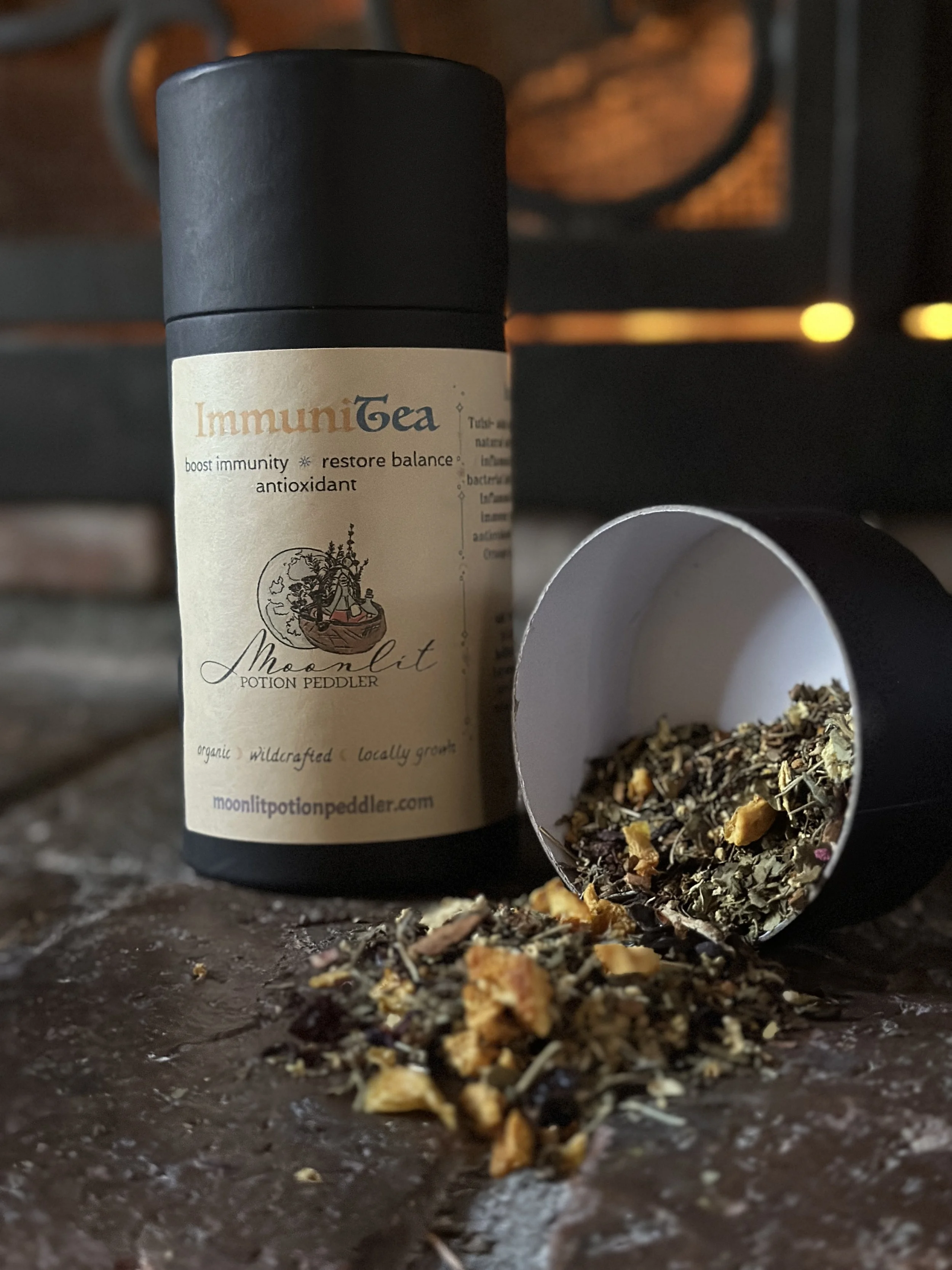 A container of ImmuniTea herbal supplement with loose herbs and dried flowers spilling out onto a dark surface. The container has a beige label with black and orange text and illustrations.
