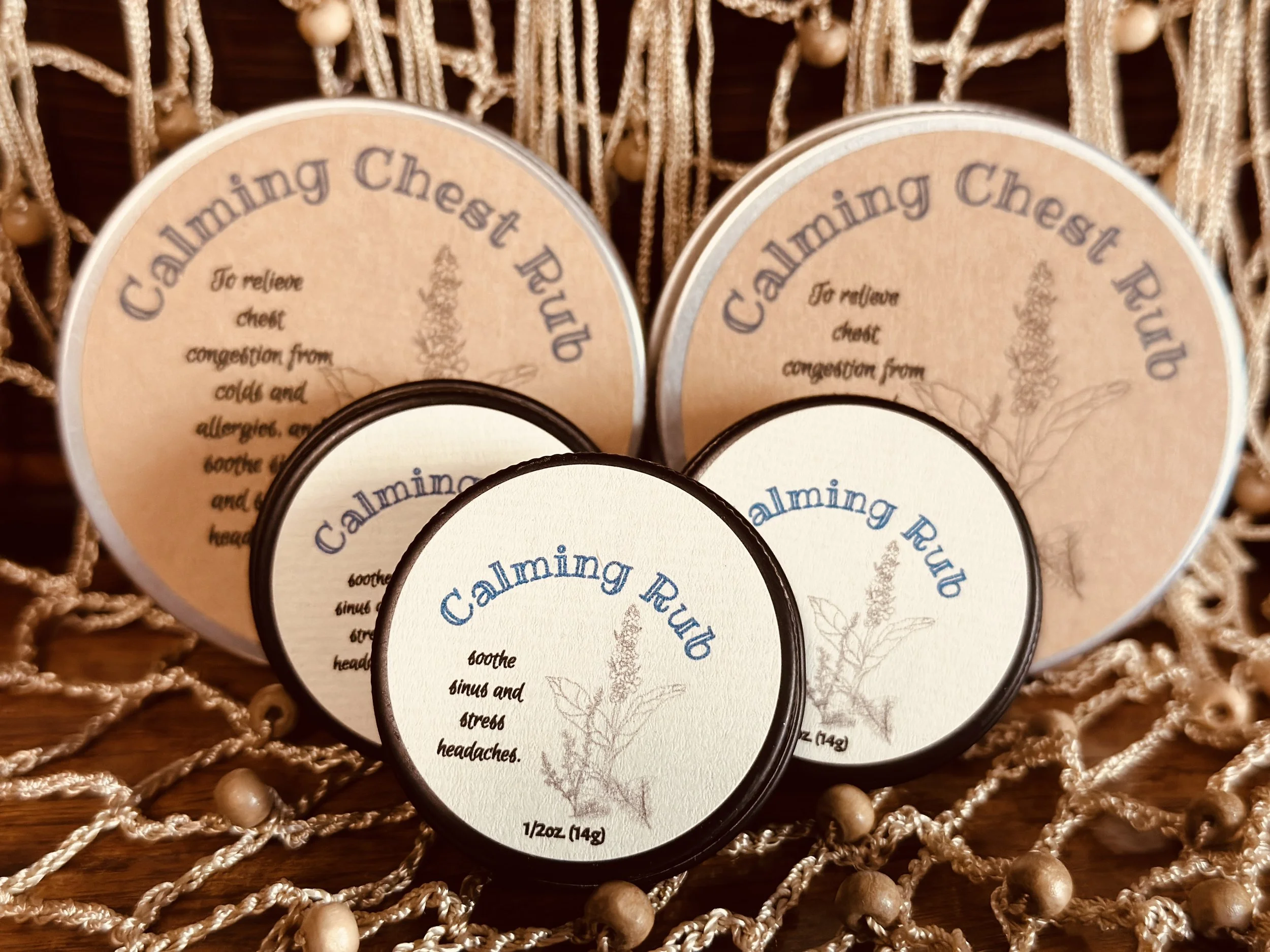 Container of calming chest rub with soothing herbal illustration, surrounded by necklaces on a woven surface.