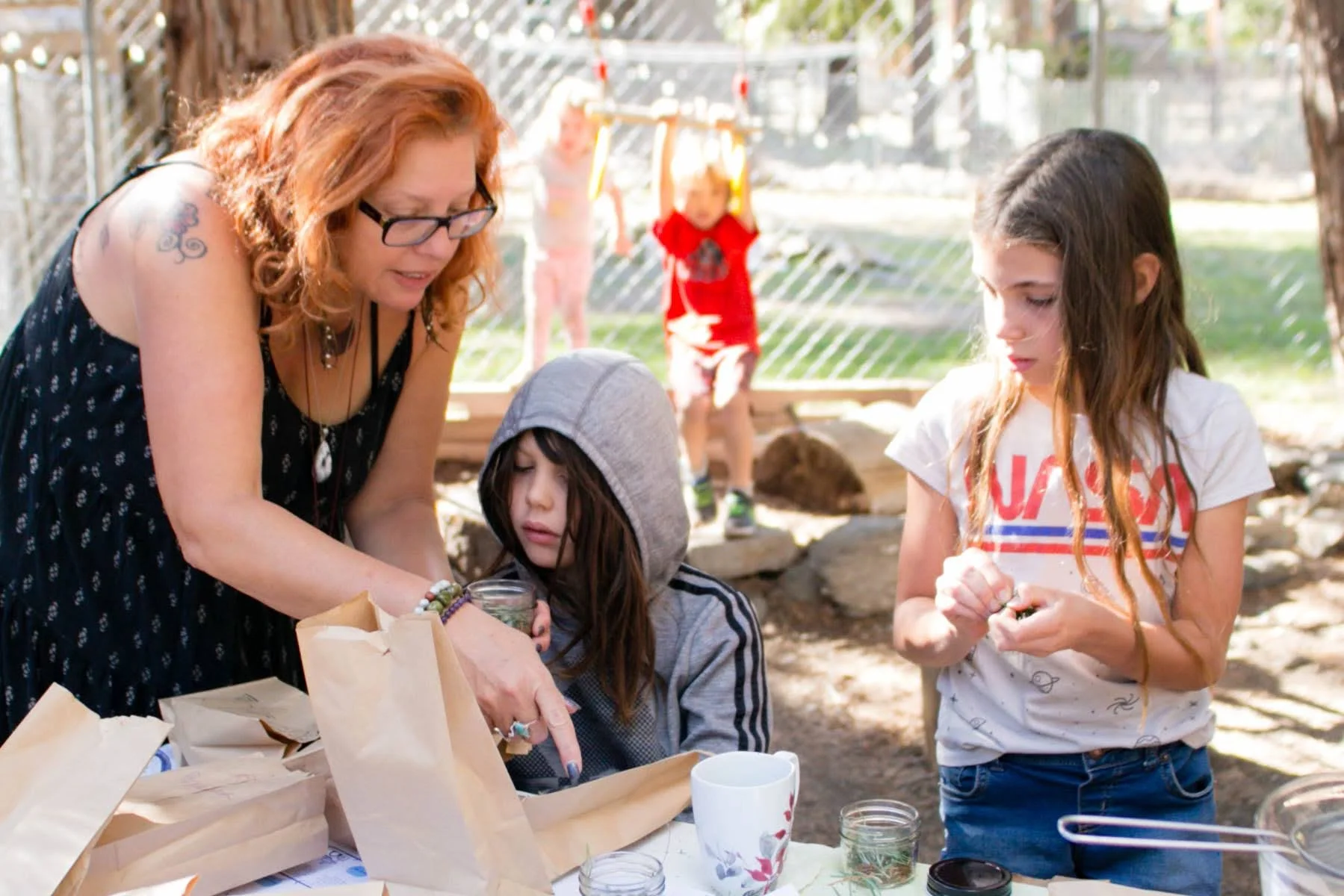Three children and a woman gathered around a table outdoors, with paper bags and cups, engaged in a creative activity, with a girl in a red shirt in the background.
