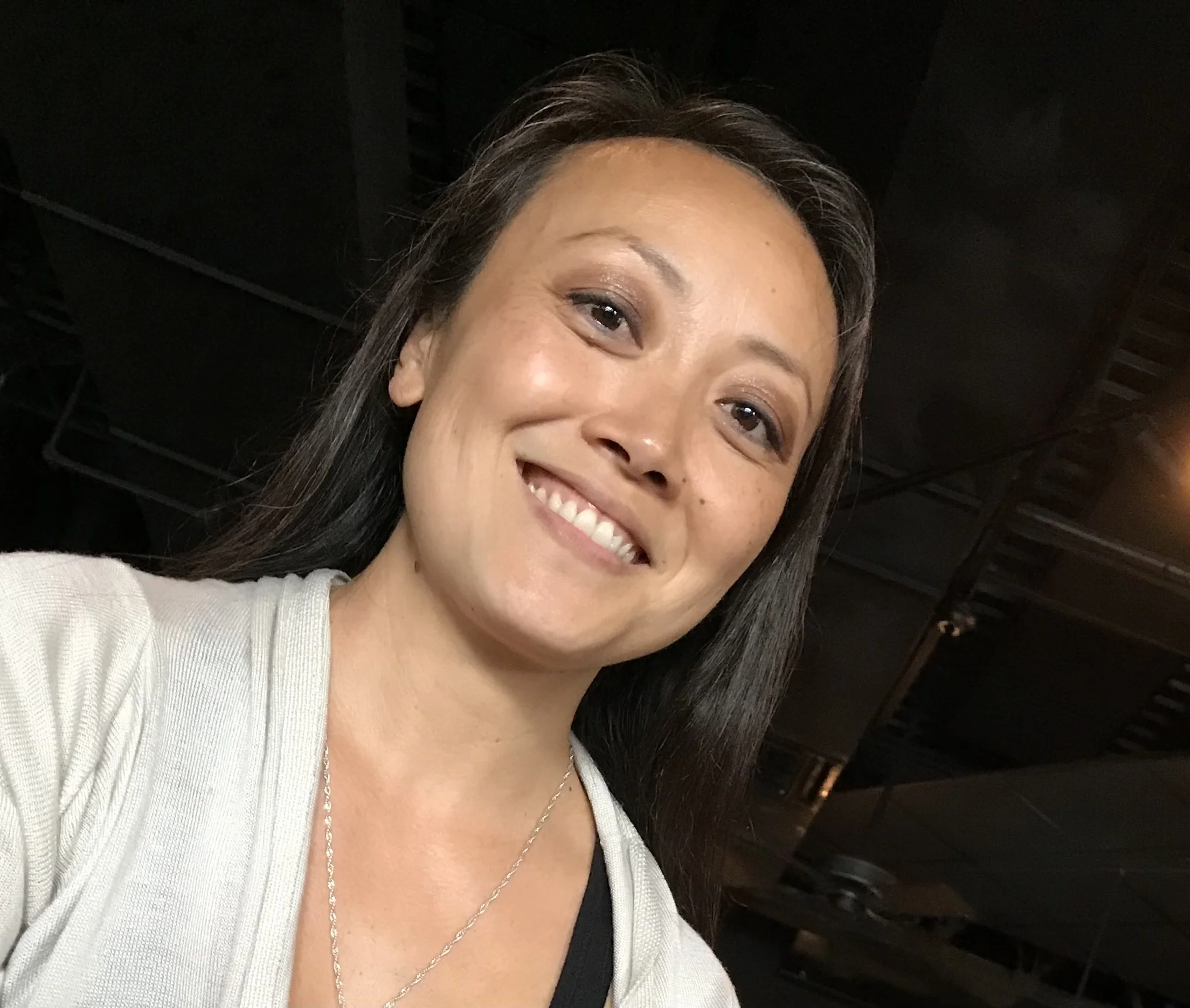 A woman with long dark hair smiling at the camera, wearing a light-colored top, jewelry, and standing indoors with a dark background.