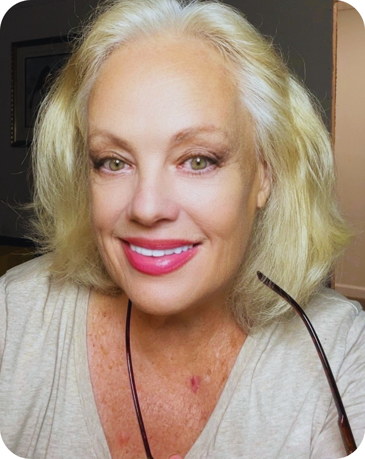 A smiling middle-aged woman with blonde hair and green eyes, wearing makeup and pink lipstick, holding a pair of glasses near her neck, in a well-lit indoor setting.