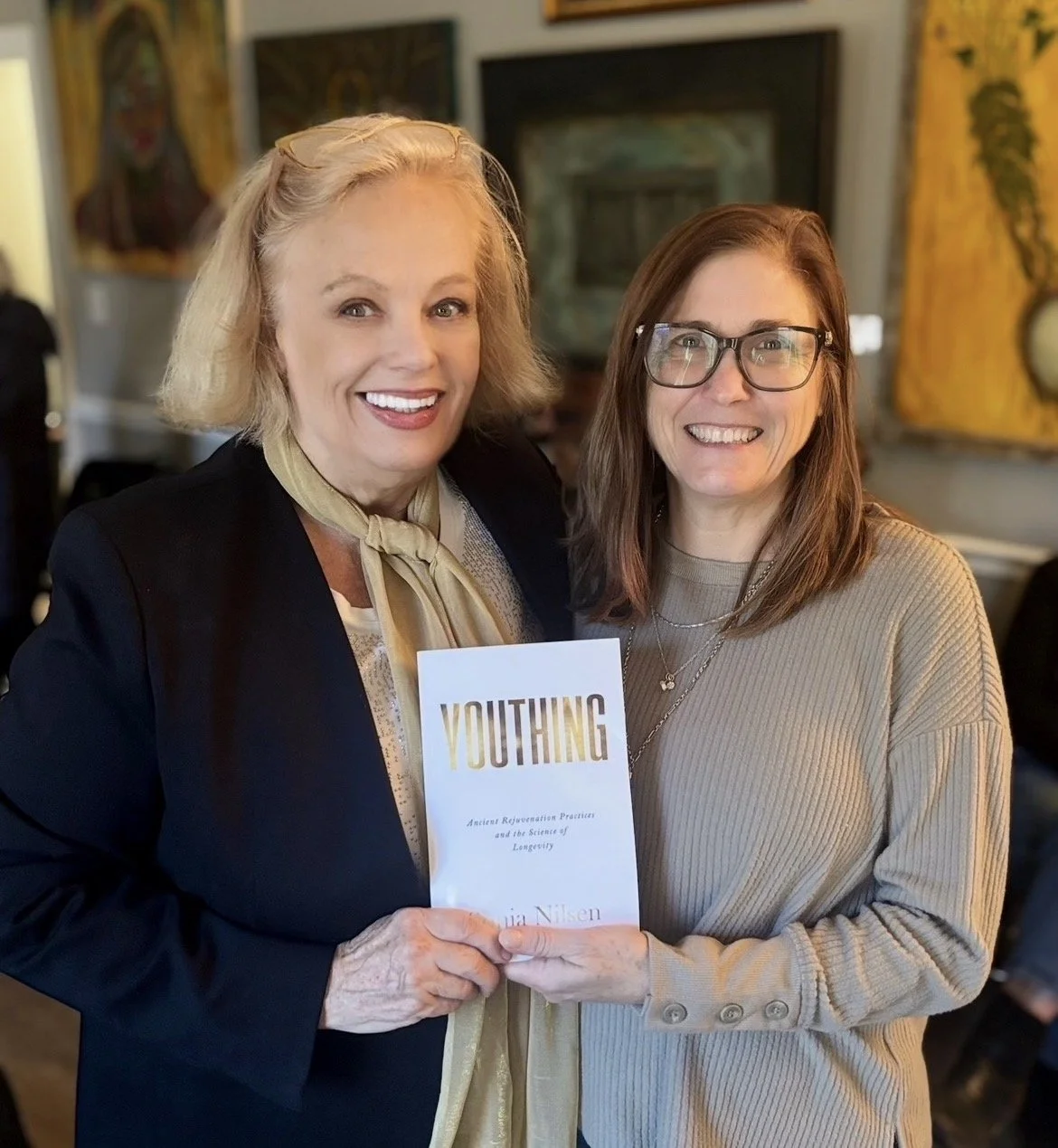Two women smiling and holding a book titled 'YOUTHING' in a room with artwork and a television in the background.