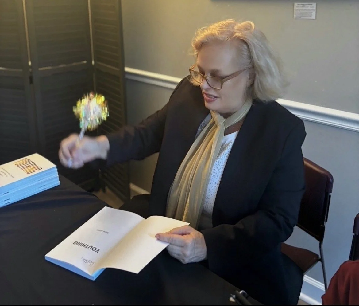 A woman with blonde hair, glasses, and a black blazer, sitting at a table, holding a book in one hand and a sparkly wand in the other, looking at the book.